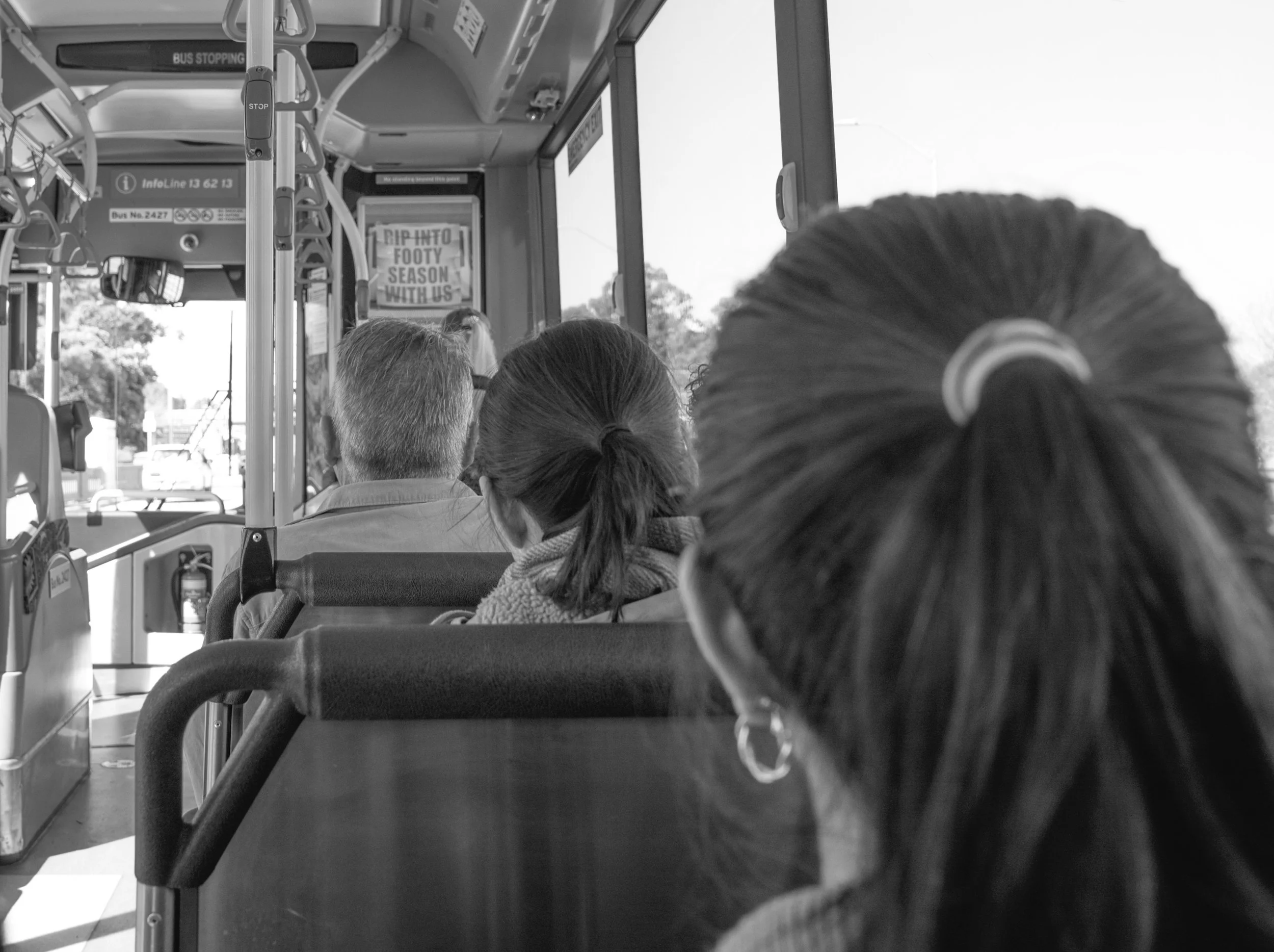 Black and white photo of people seated on a bus, viewed from the back, with a person in the foreground having dark hair in a ponytail. Others are seated facing forward, and a window on the right reveals trees and sky outside.