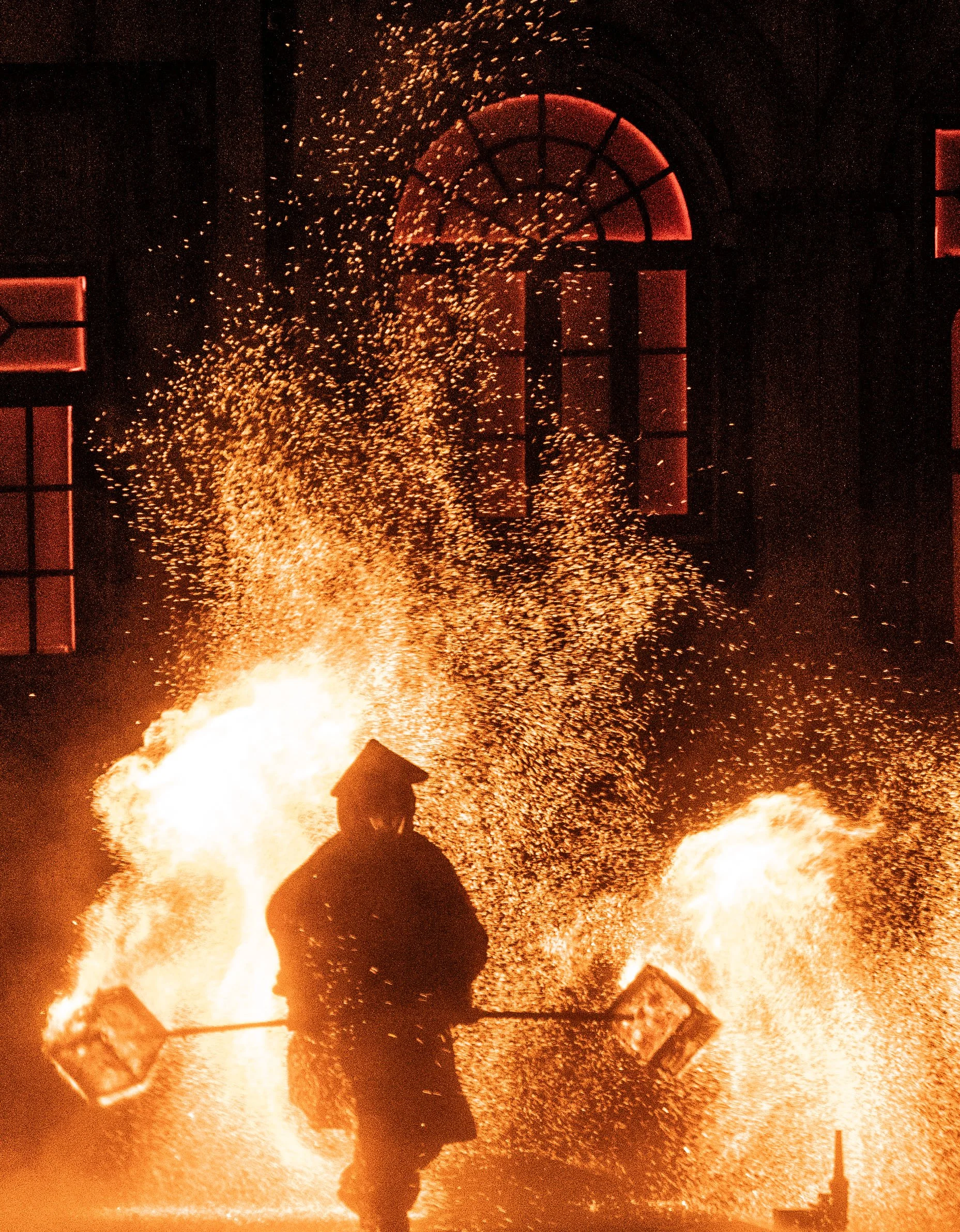 A person working with a flaming forge or pyrotechnic display at night, with sparks flying and glowing windows in the background.