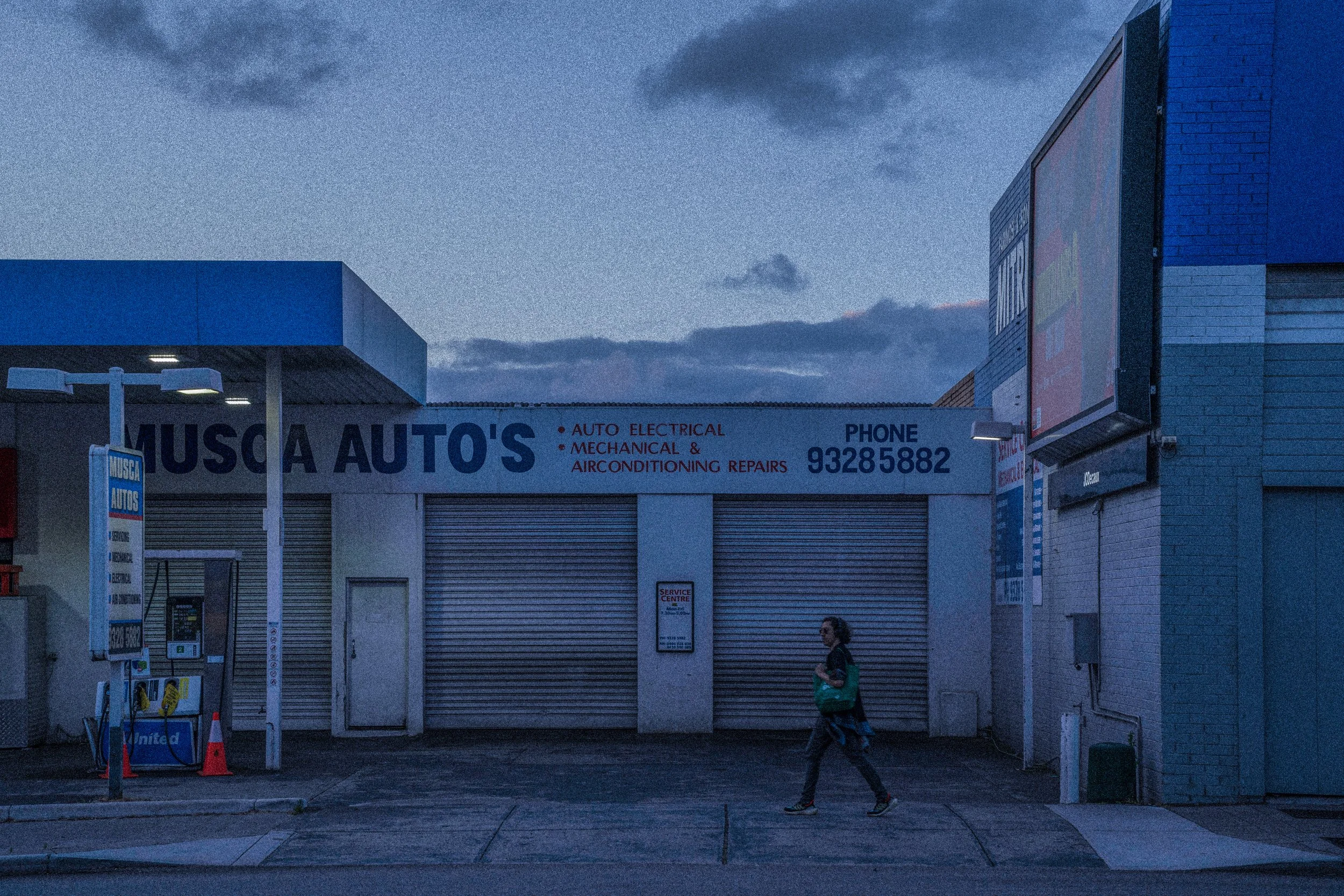 A building with a sign that reads 'Musca Auto's' and mentions auto electrical, mechanical, and air conditioning repairs, with a phone number '93285882'. There are closed roller shutters, a woman walking on the sidewalk, and a blue sky with some cloud