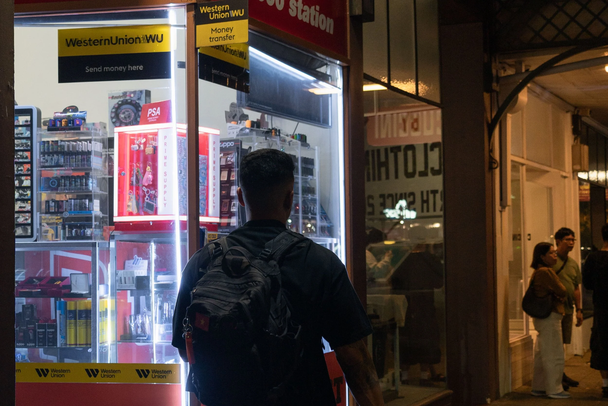 A person with a backpack standing outside a store with a brightly lit display case containing various electronic accessories, at night. Other people are standing nearby outside the store.