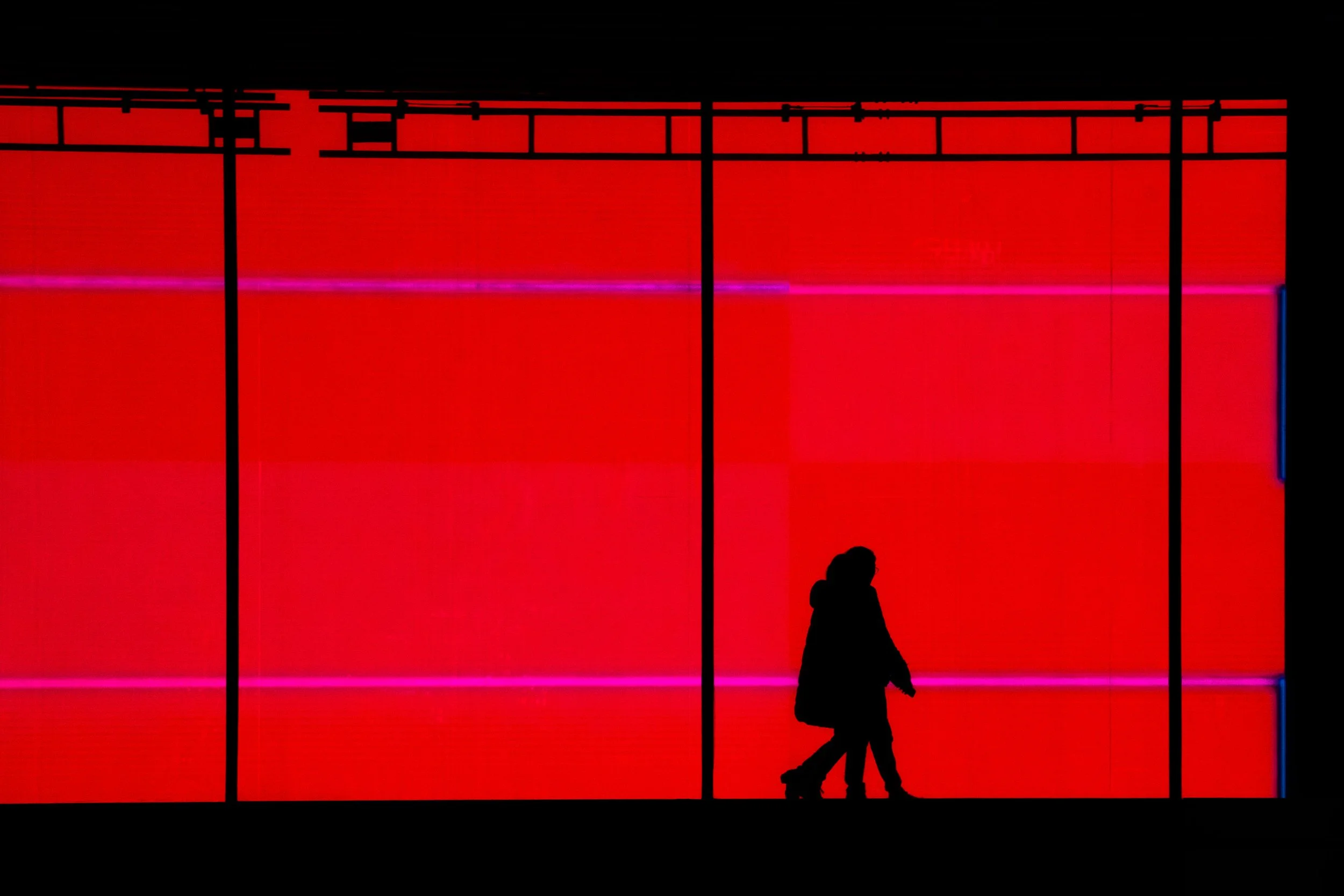 Silhouette of a person walking past a vibrant red neon-lit wall with horizontal purple and pink light streaks.