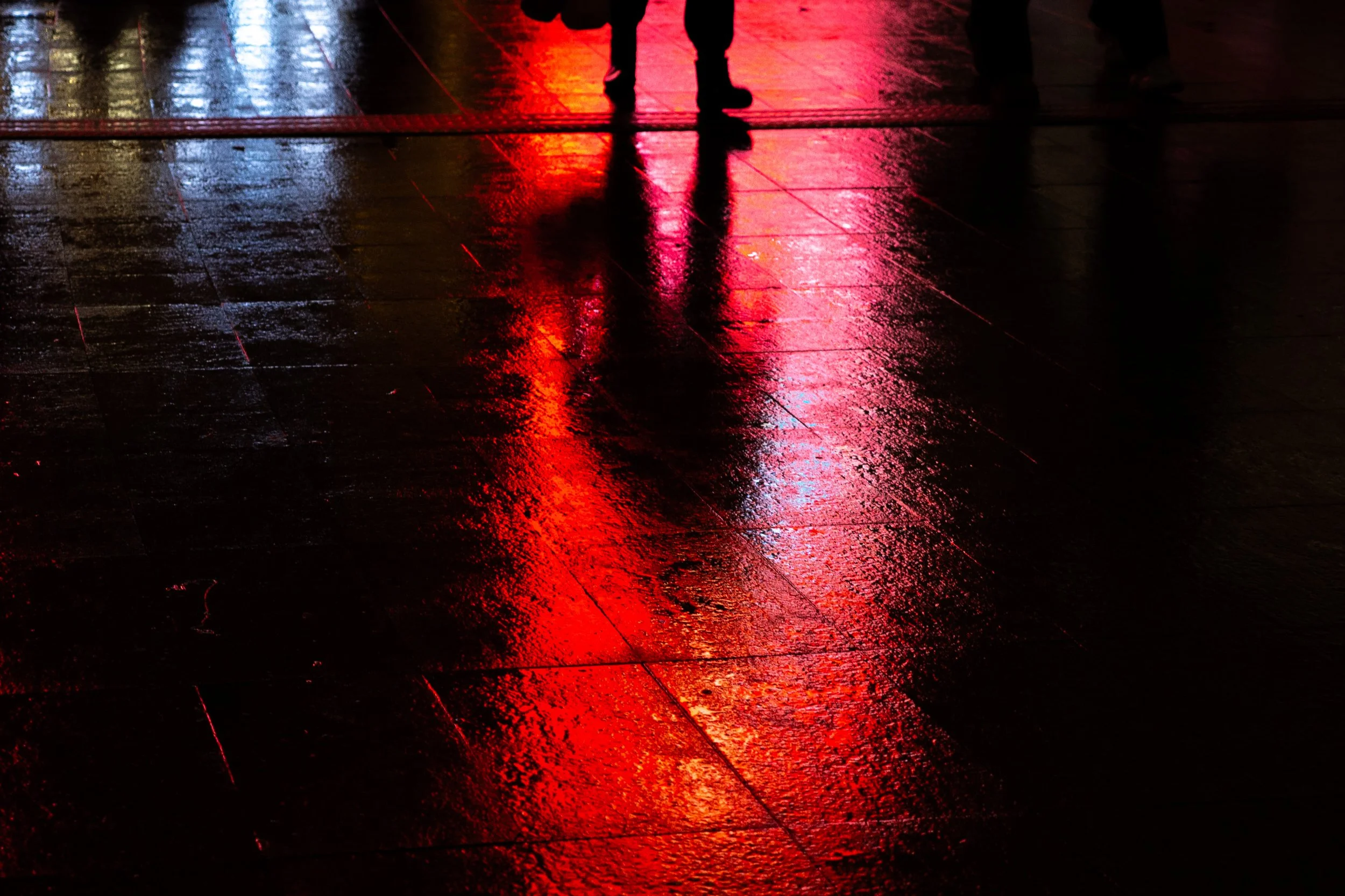 Silhouette of a person walking on a wet floor reflecting red and yellow city lights at night.