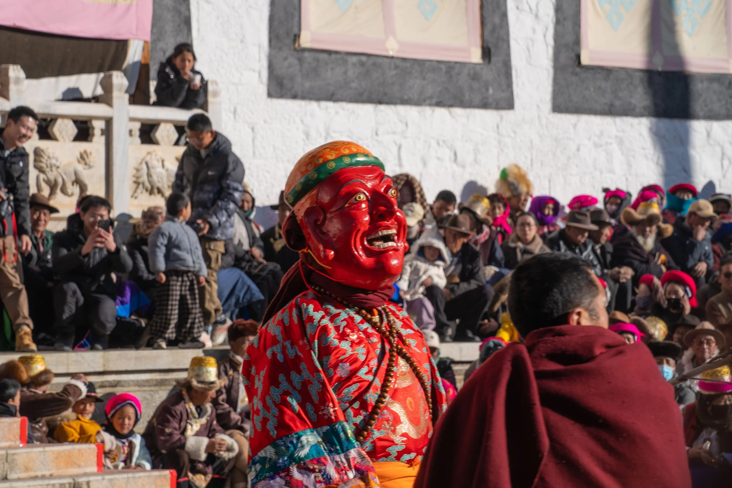 A person wearing a red theatrical mask and colorful clothing participating in a cultural festival, with a crowd of onlookers in warm clothing and traditional hats watching in the background.