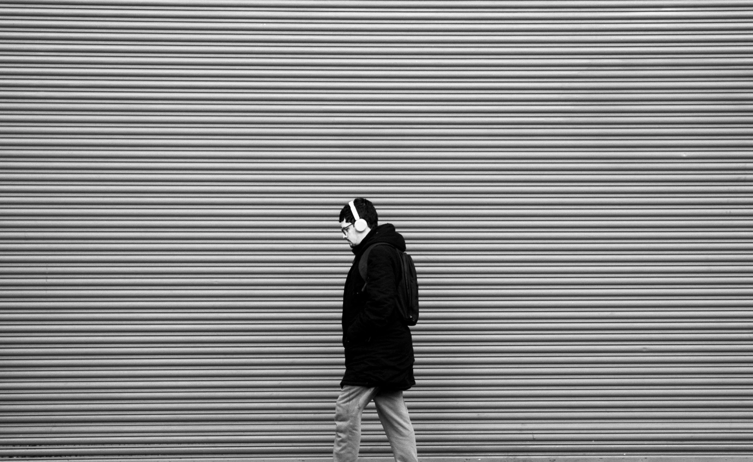 A man wearing headphones, glasses, a dark coat, and light-colored pants walking past a large, closed rolling metal shutter.