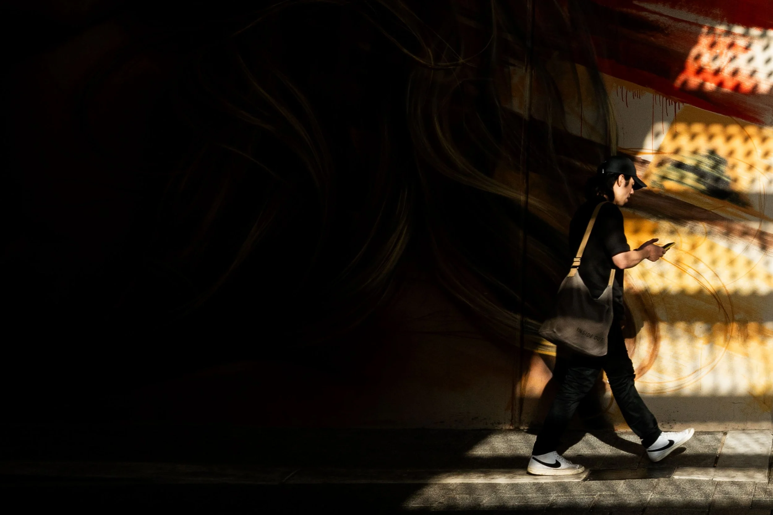 A person in black clothing with a black cap, white sneakers, and a black shoulder bag walking and looking at their phone, with a colorful mural on the wall behind them.