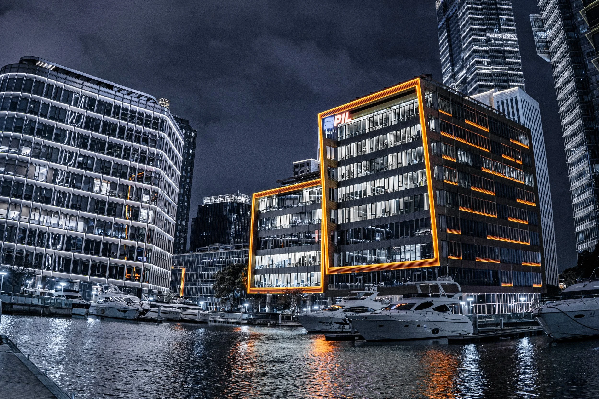 Nighttime view of modern city buildings with boats docked on a canal, illuminated windows, and a building with orange neon lighting and the 'PIL' sign.