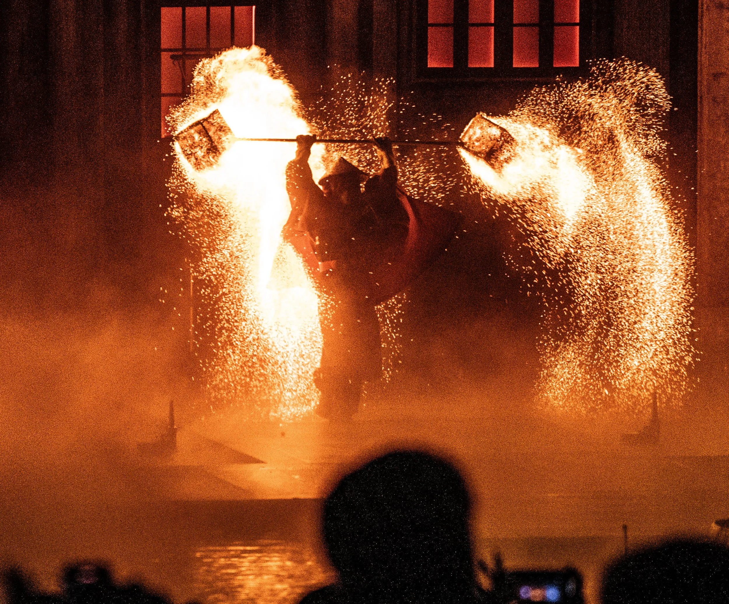 A performer shooting fire with two torches in a dark indoor setting, with sparks flying around.