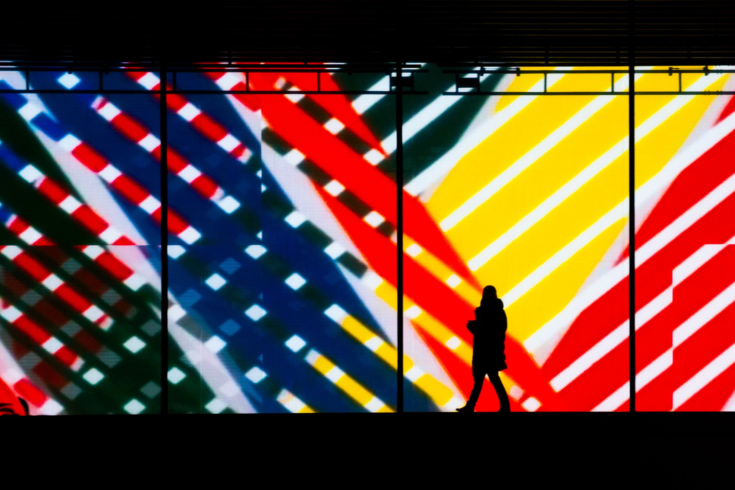 Silhouette of a person walking past a large colorful digital display with diagonal red, blue, yellow, and white lines and patterns.