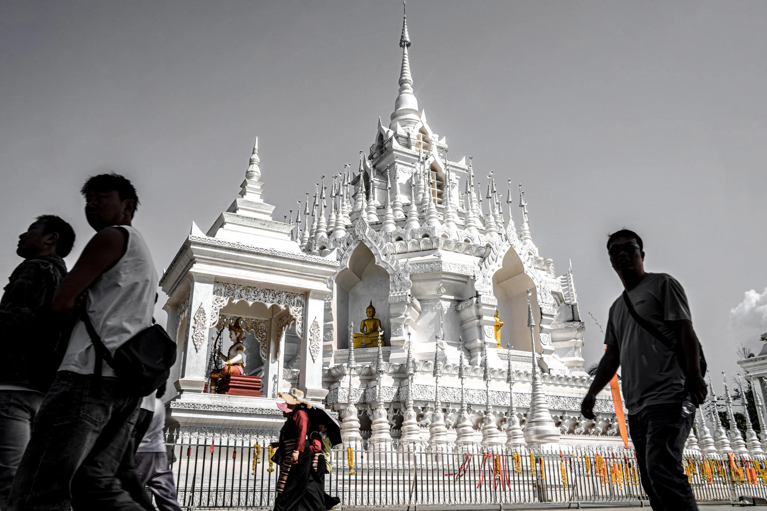A white ornate temple with golden Buddha statues on its steps, surrounded by a black fence, and several people walking in front of it.