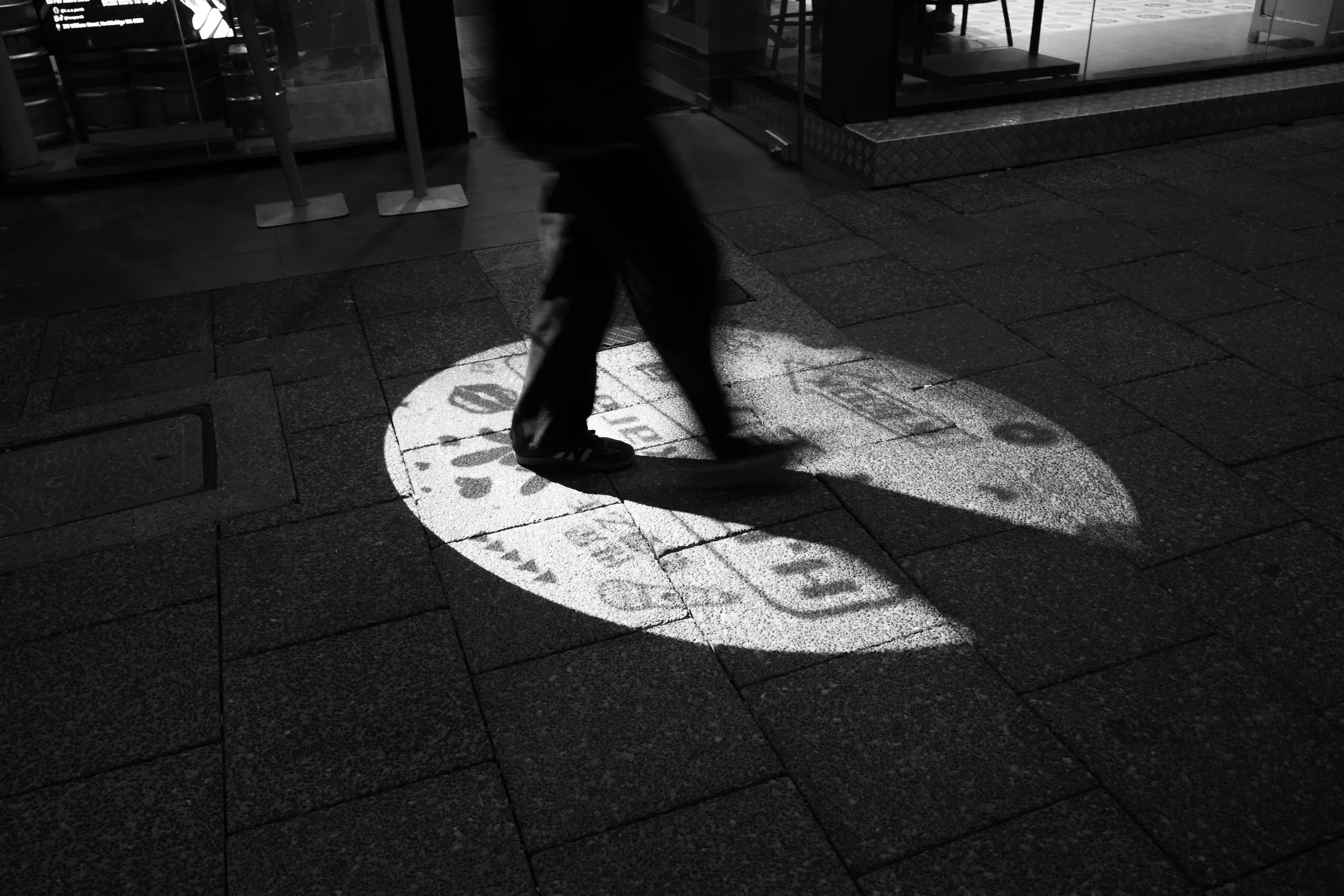 A person walking on a sidewalk at night, with a cone-shaped projection of text and graphics on the ground from a nearby storefront window.