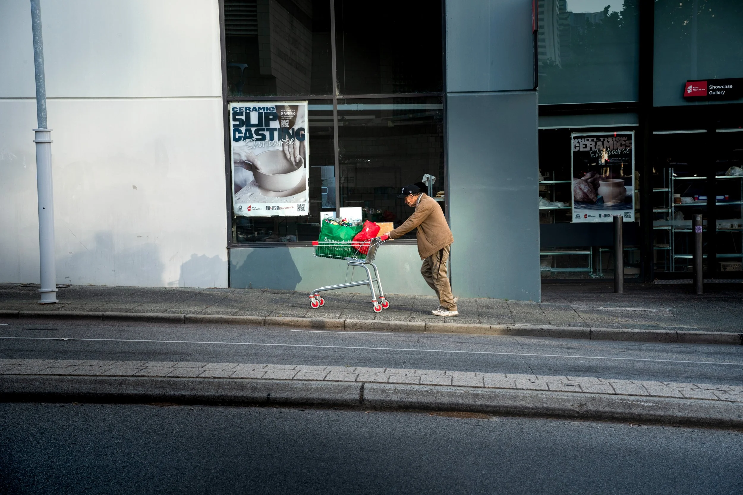 A man wearing a brown jacket and cap pushing a shopping cart on a city sidewalk in front of a glass building