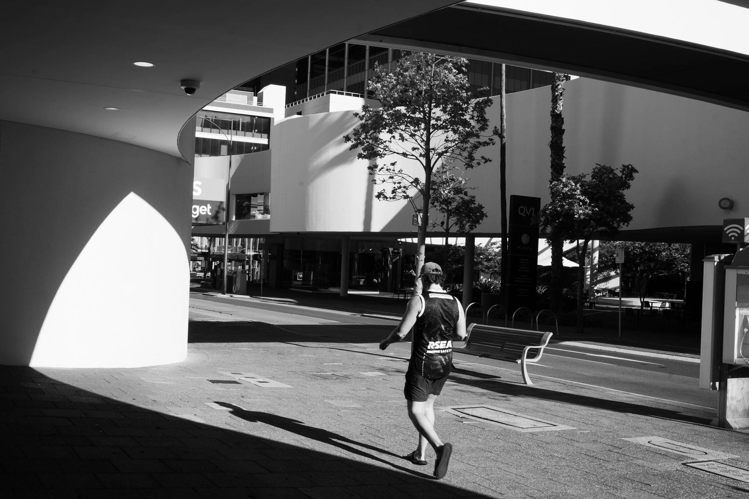 A person jogging outdoors in an urban area with modern buildings, trees, benches, and a parking lot in the background. Black and white photo.