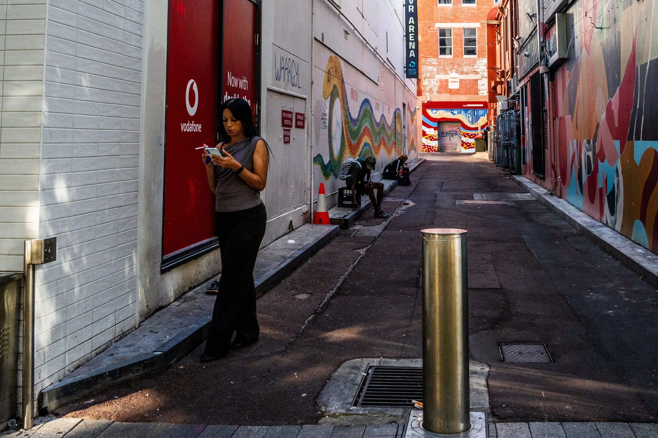 A woman with long dark hair, wearing a gray sleeveless top and black pants, stands on a city sidewalk looking at her phone. In the background, two people sit on benches next to a mural of a colorful, abstract snake on the wall, under a partly sunny s