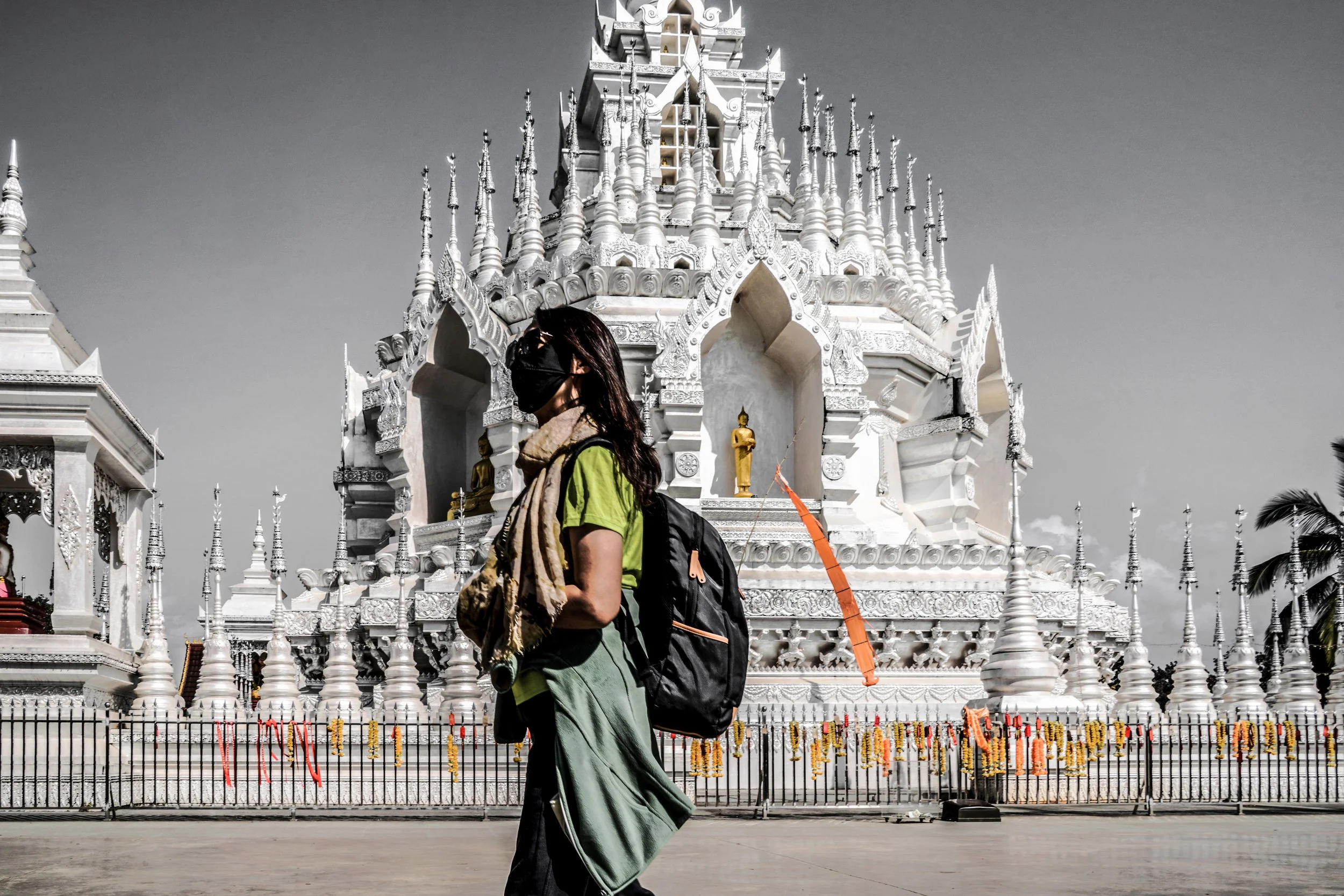 A person wearing a black mask, green shirt, and scarf around the neck, carrying a black backpack, walking past a large, ornate white temple with gold statues in the background.