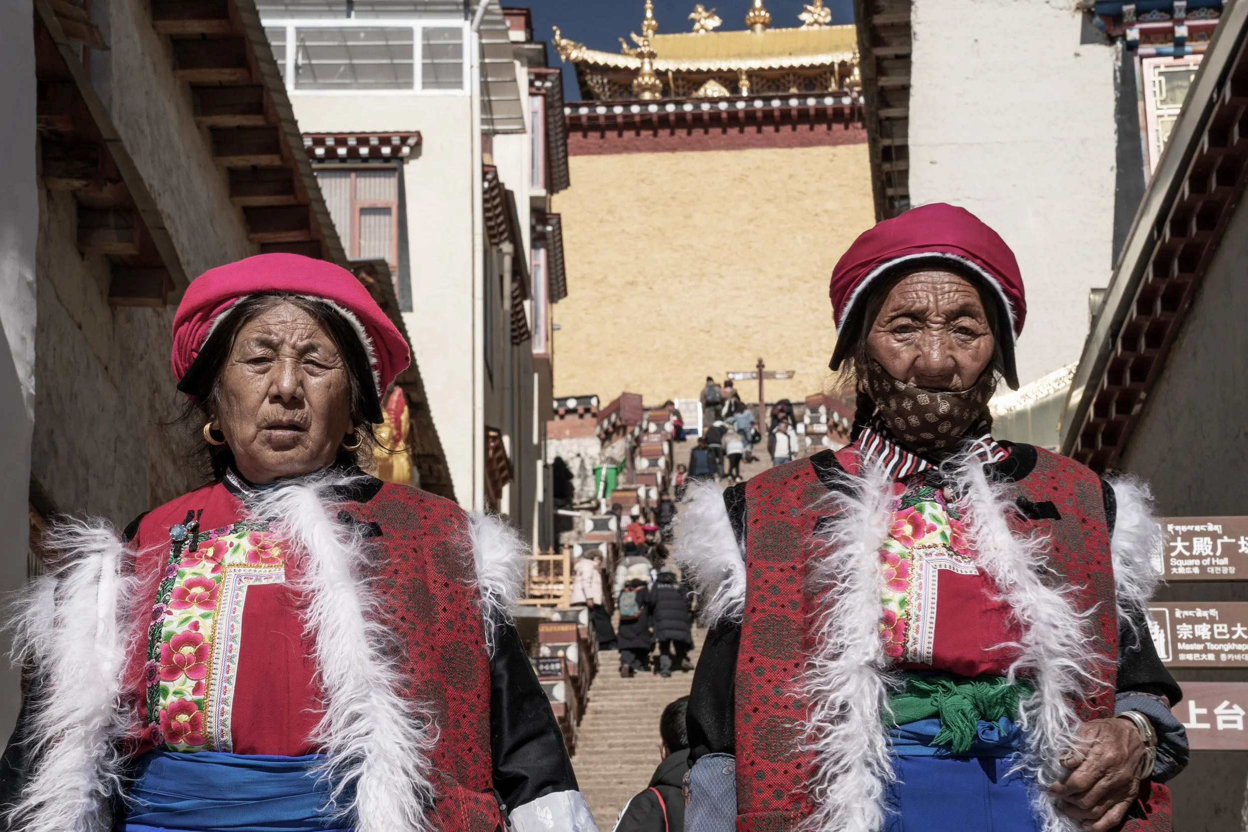 Two elderly women dressed in traditional Tibetan clothing standing in front of a large staircase leading to a temple or monastery with Tibetan script signboards nearby.
