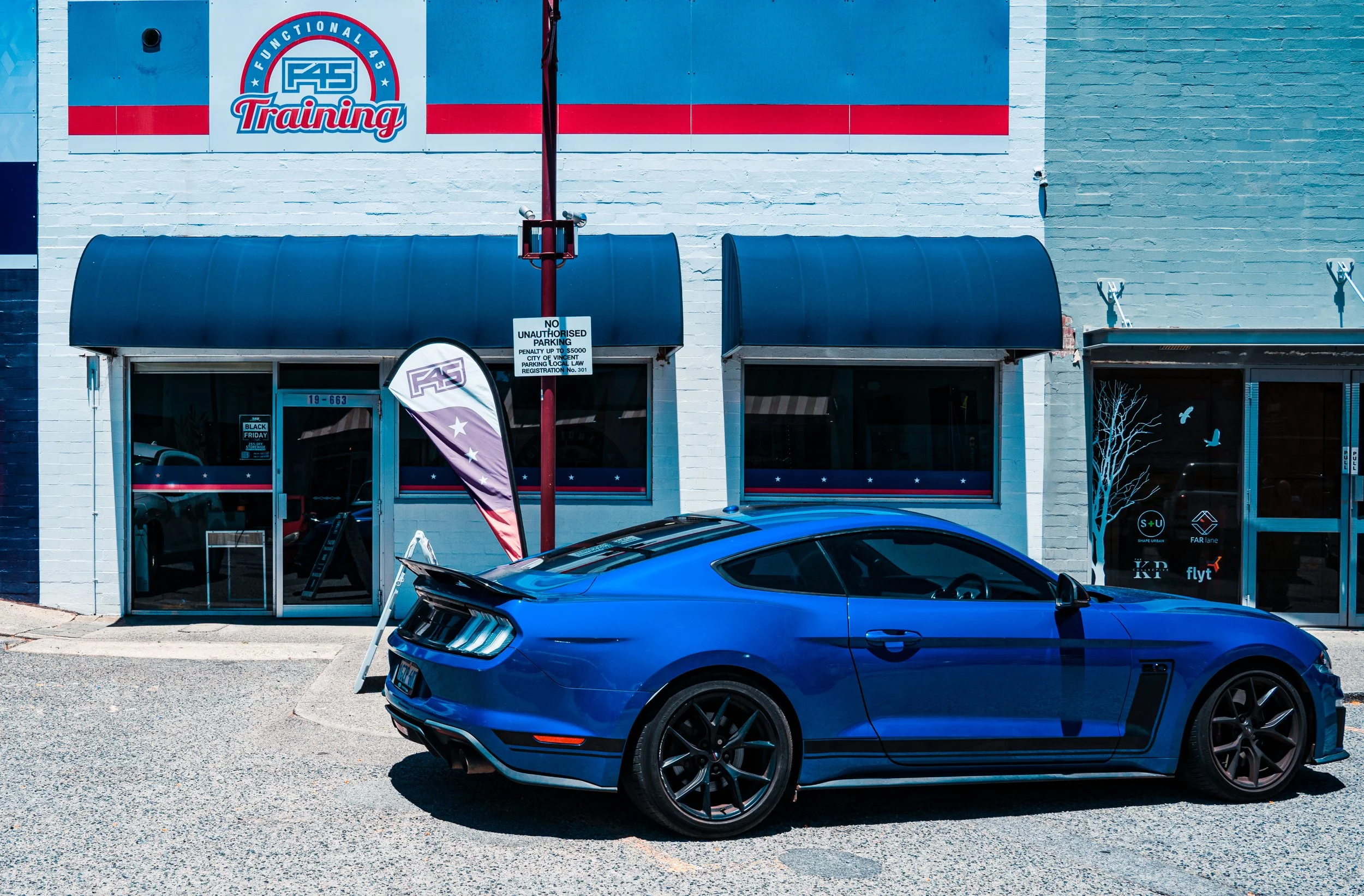 A blue Ford Mustang parked outside a training facility with an American flag flag and signage in the background.