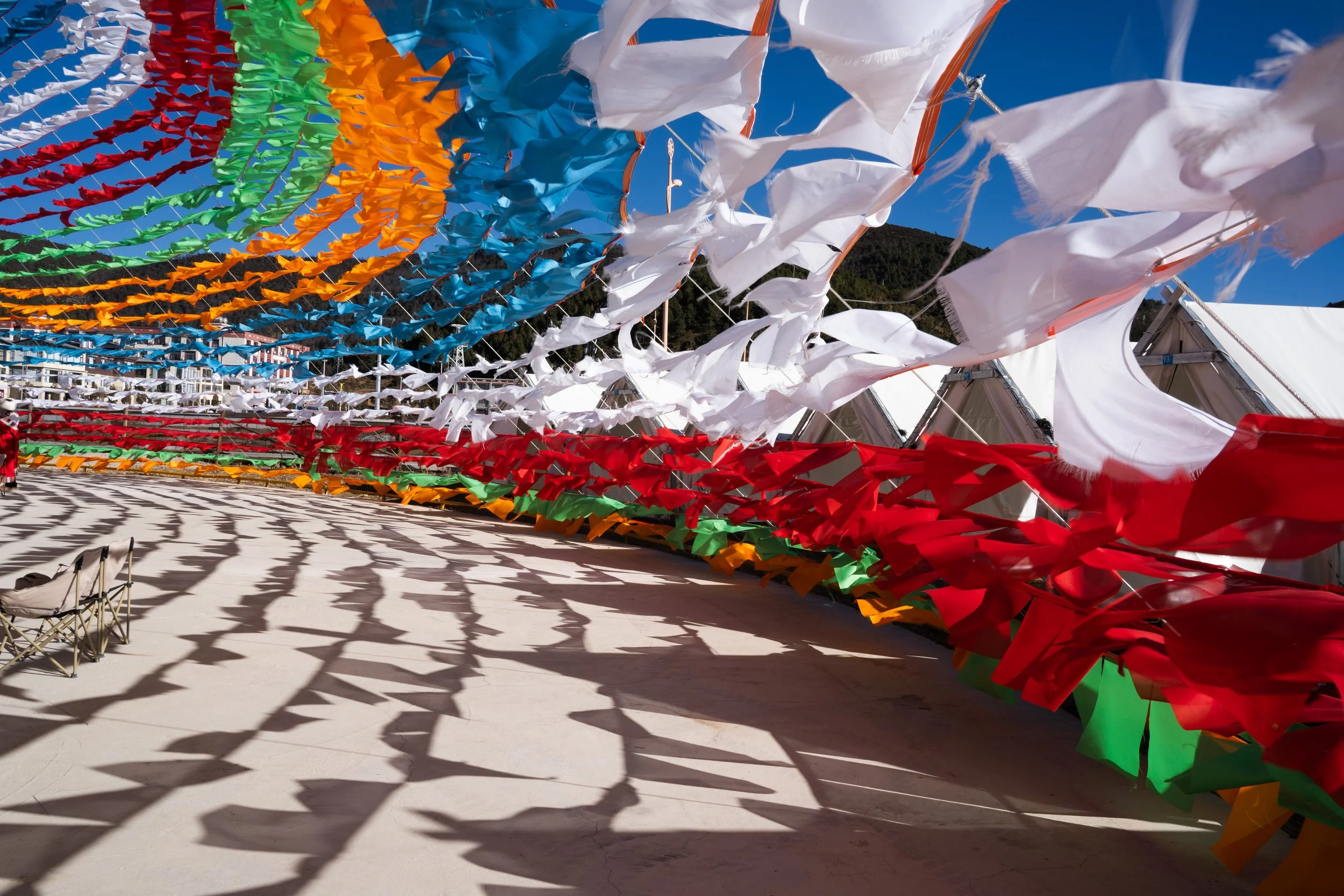 Colorful fabric pennants and white flags flutter in the wind, casting shadow patterns on the ground, against a clear blue sky with mountains in the background.