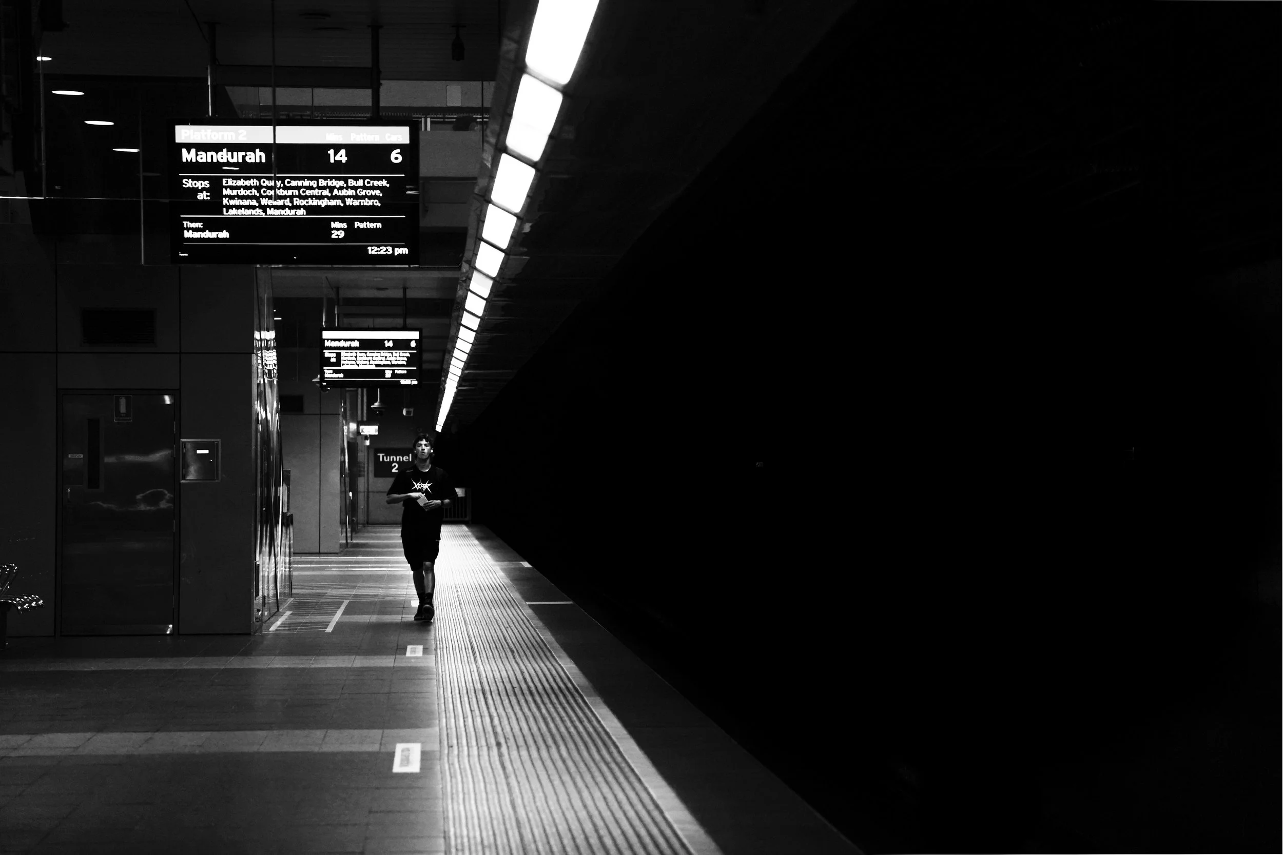 A person walking in a train station with digital signs displaying train information. The station has a dark, modern interior, with illuminated signage and a spacious platform.