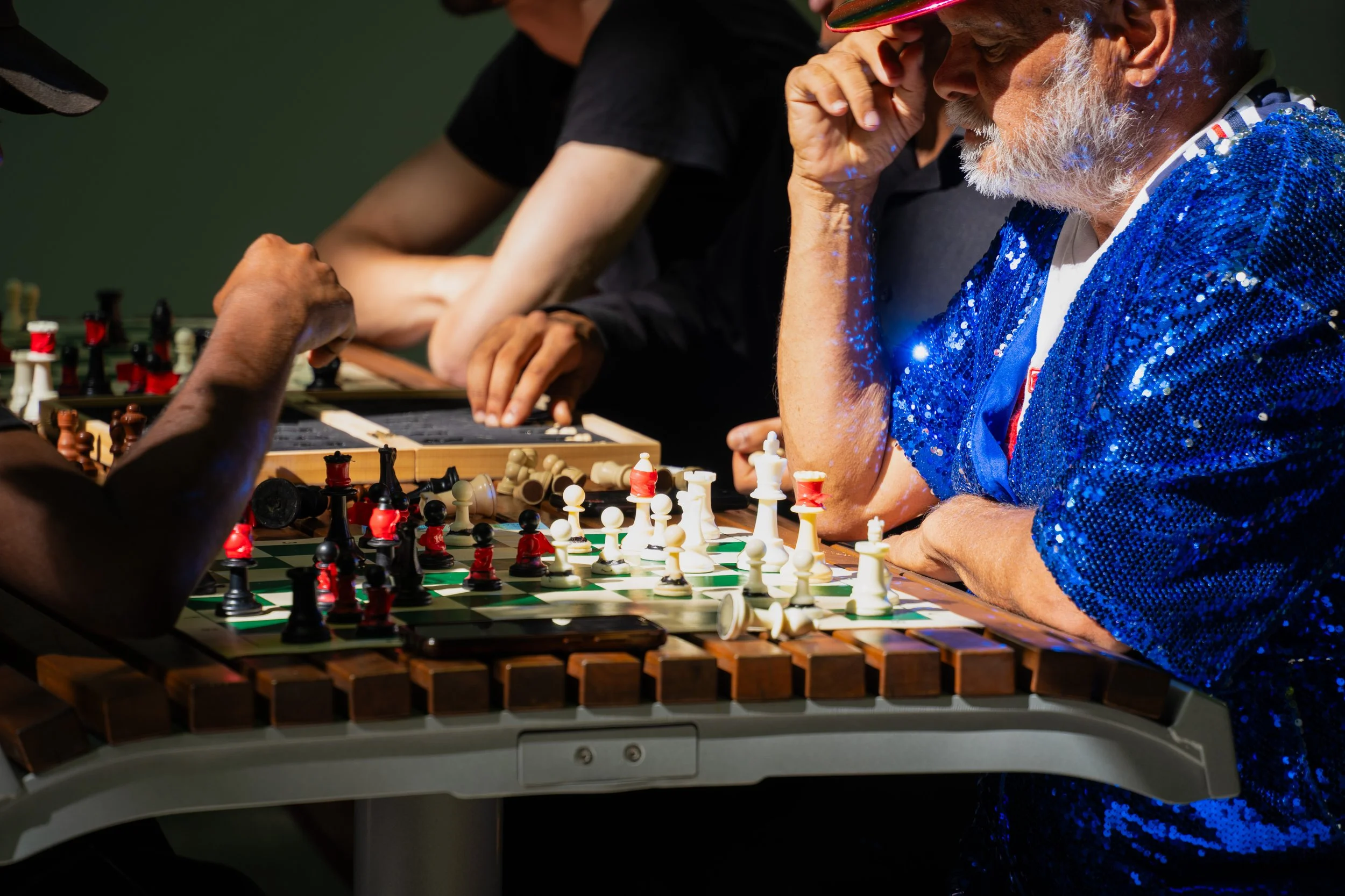People playing chess at a table, with one man wearing a blue sequined jacket and a cap, and others concentrating on the game.