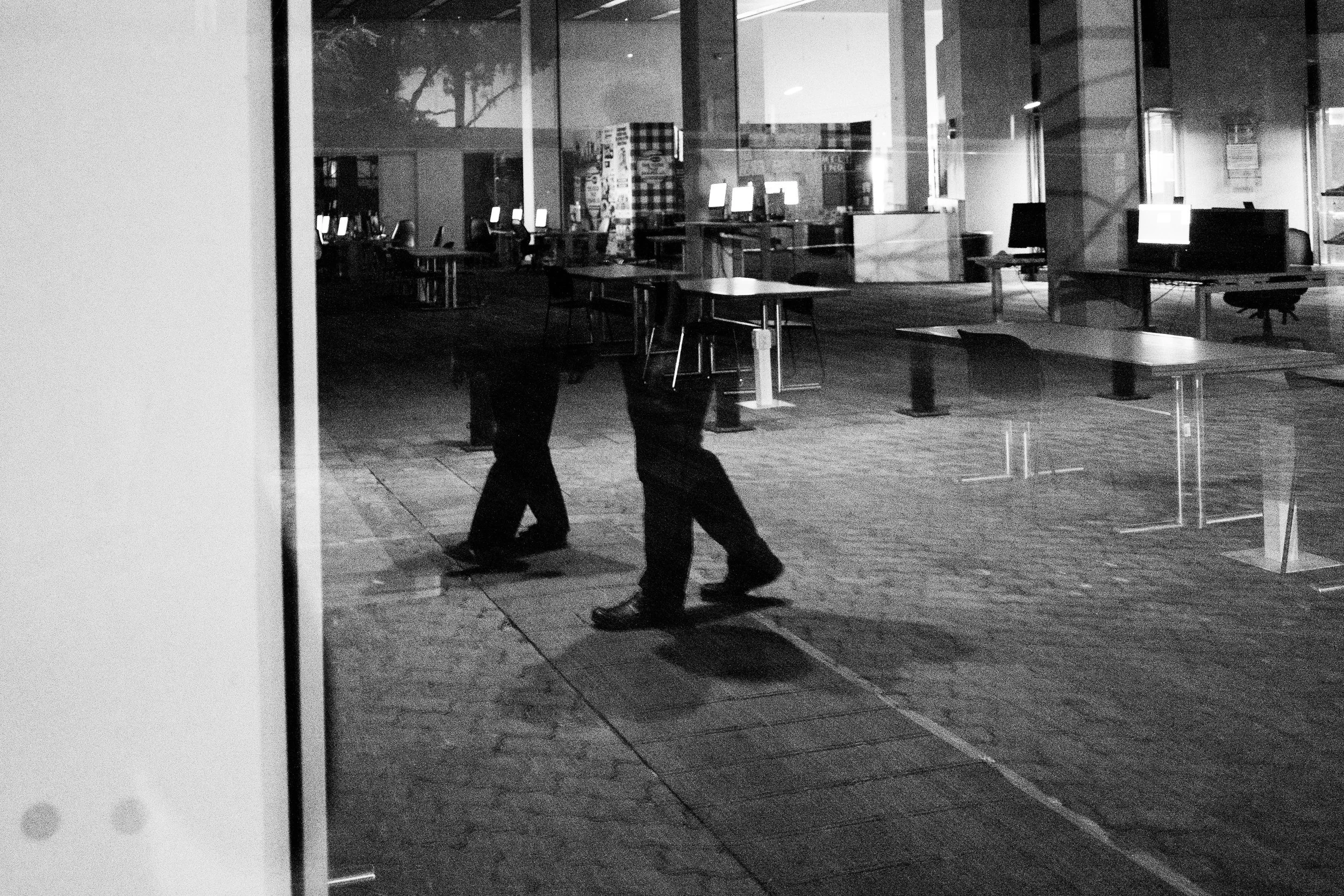 Reflection of two walking people seen through a glass window, with an empty computer lab or office space inside in the background.