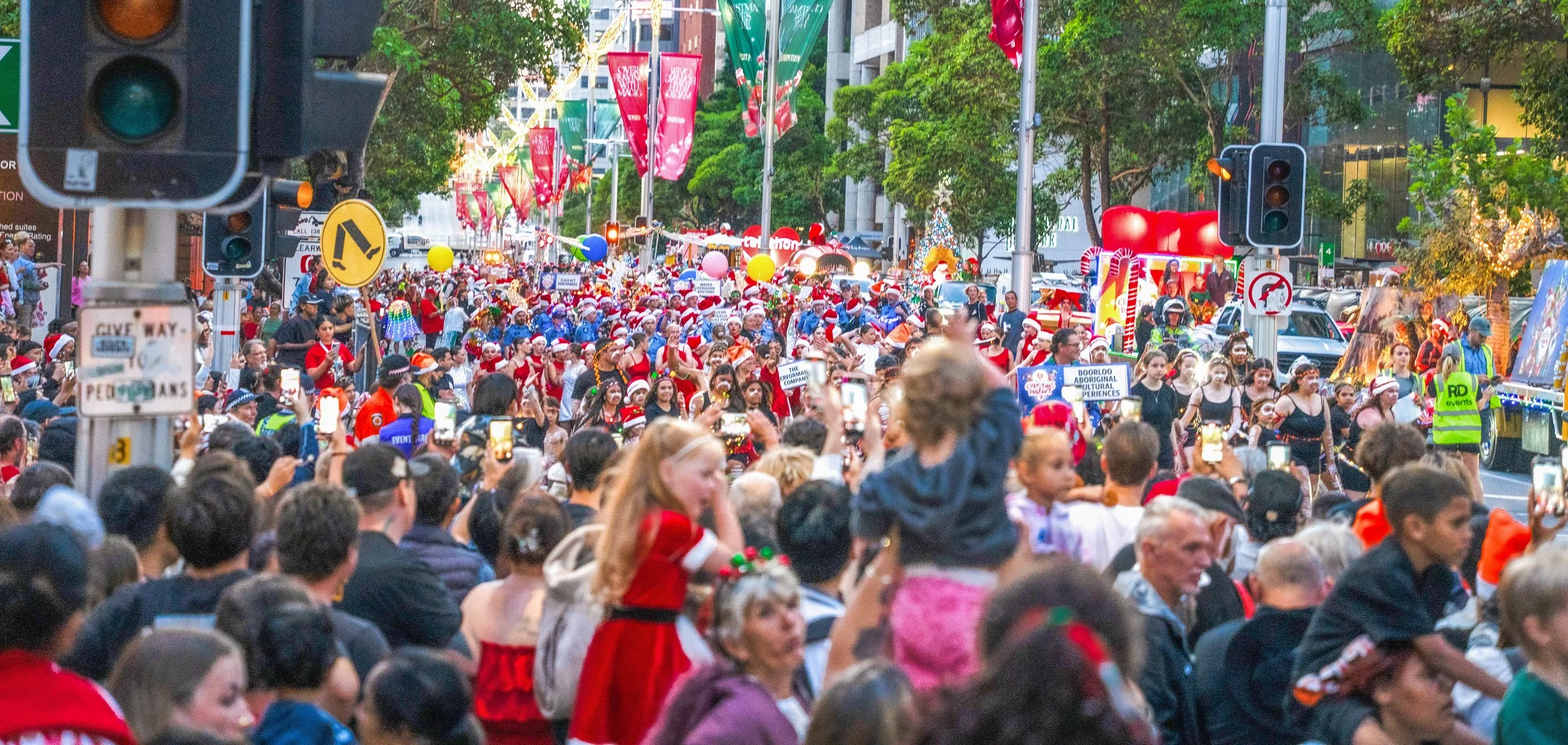 Crowd of people participating in a festive parade or event on a city street, many wearing Christmas hats, with balloons and holiday decorations, and traffic lights visible in the background.