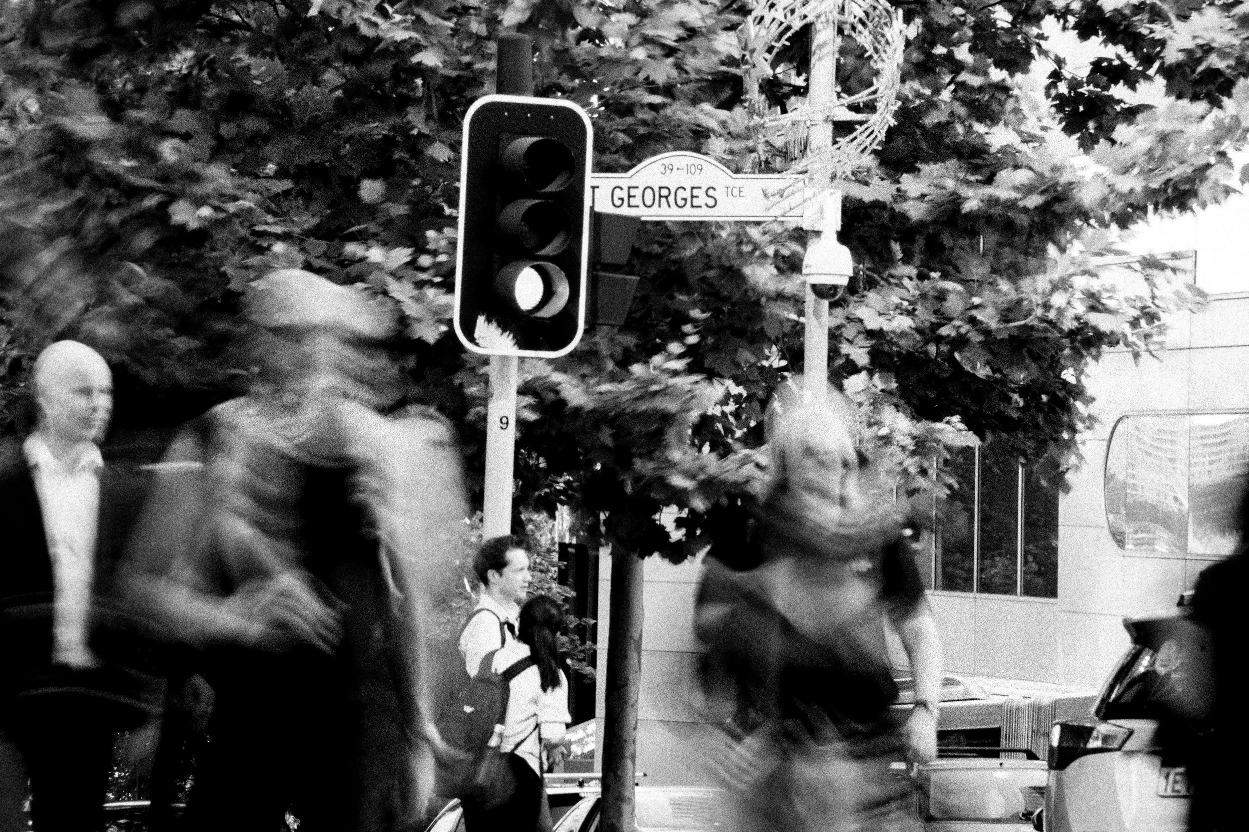Black and white photo of pedestrians crossing the street at a busy city intersection with a traffic light and street sign that reads 'Gorges'.