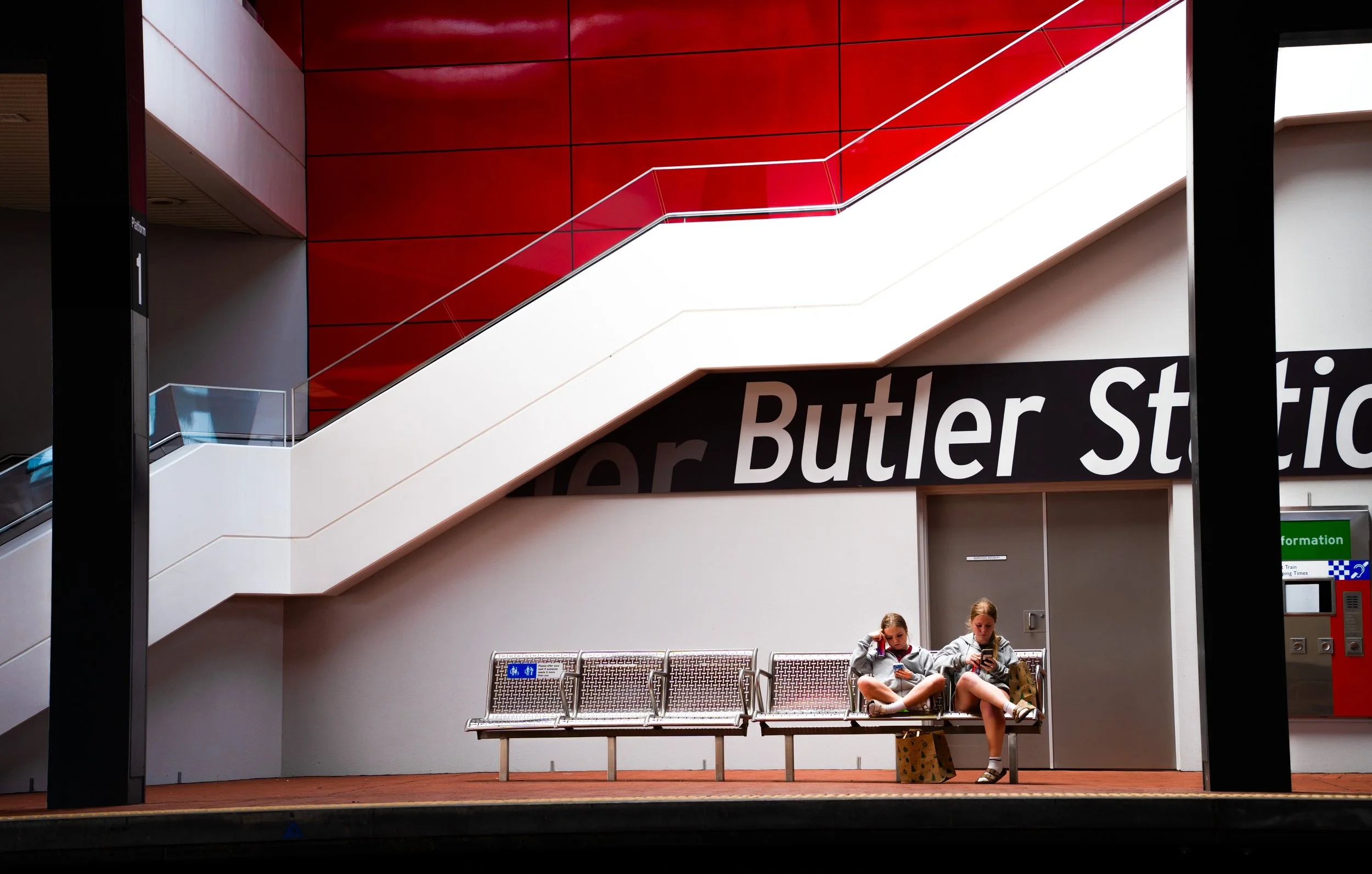 Two young women sit on a bench at Butler Station, one talking on her phone and the other looking at her phone, with a suitcase beside them, in a modern train station with red and white walls and an escalator.