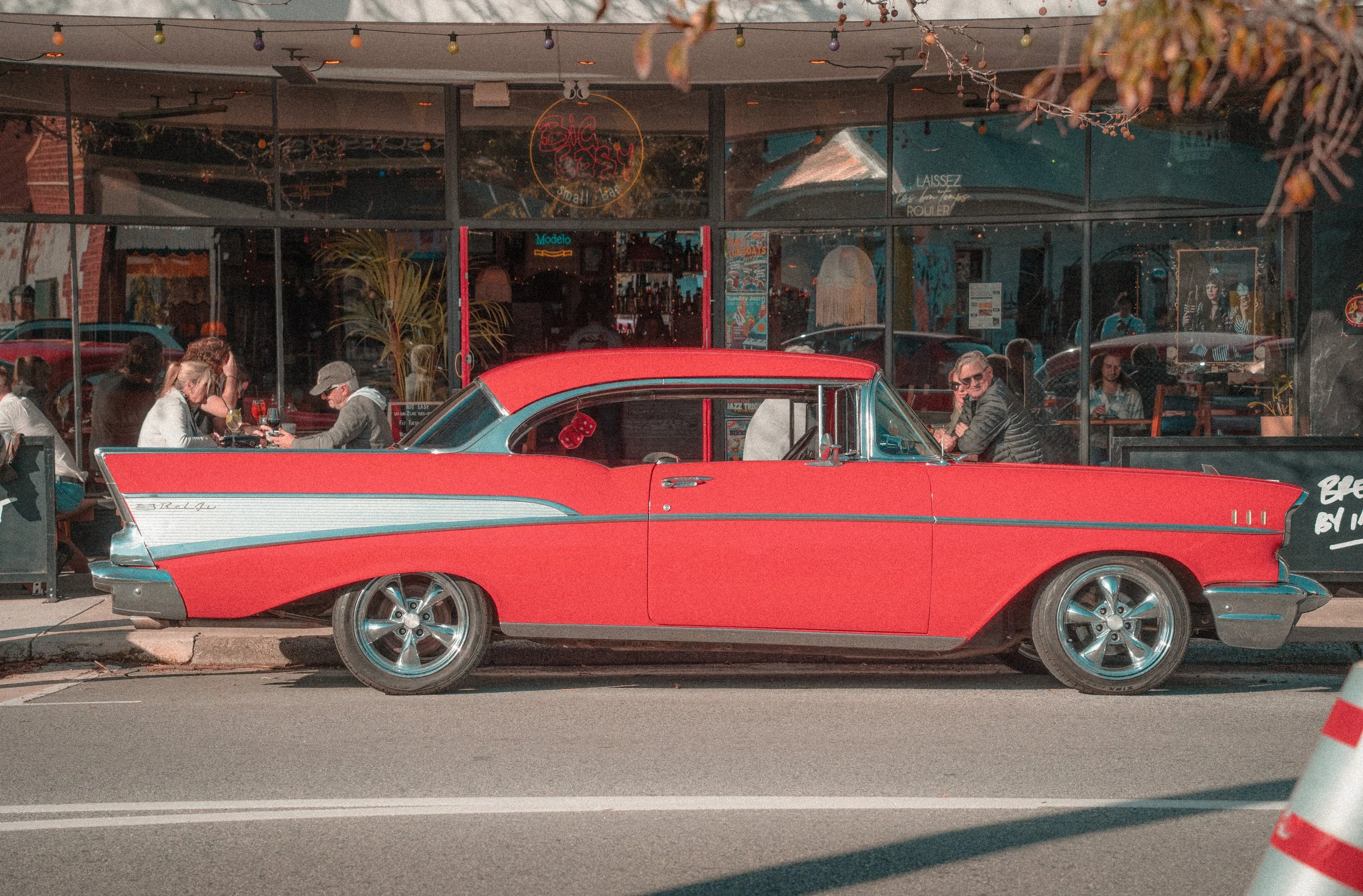 A red vintage car parked on the side of the street outside a restaurant, with people dining inside visible through the large glass windows.