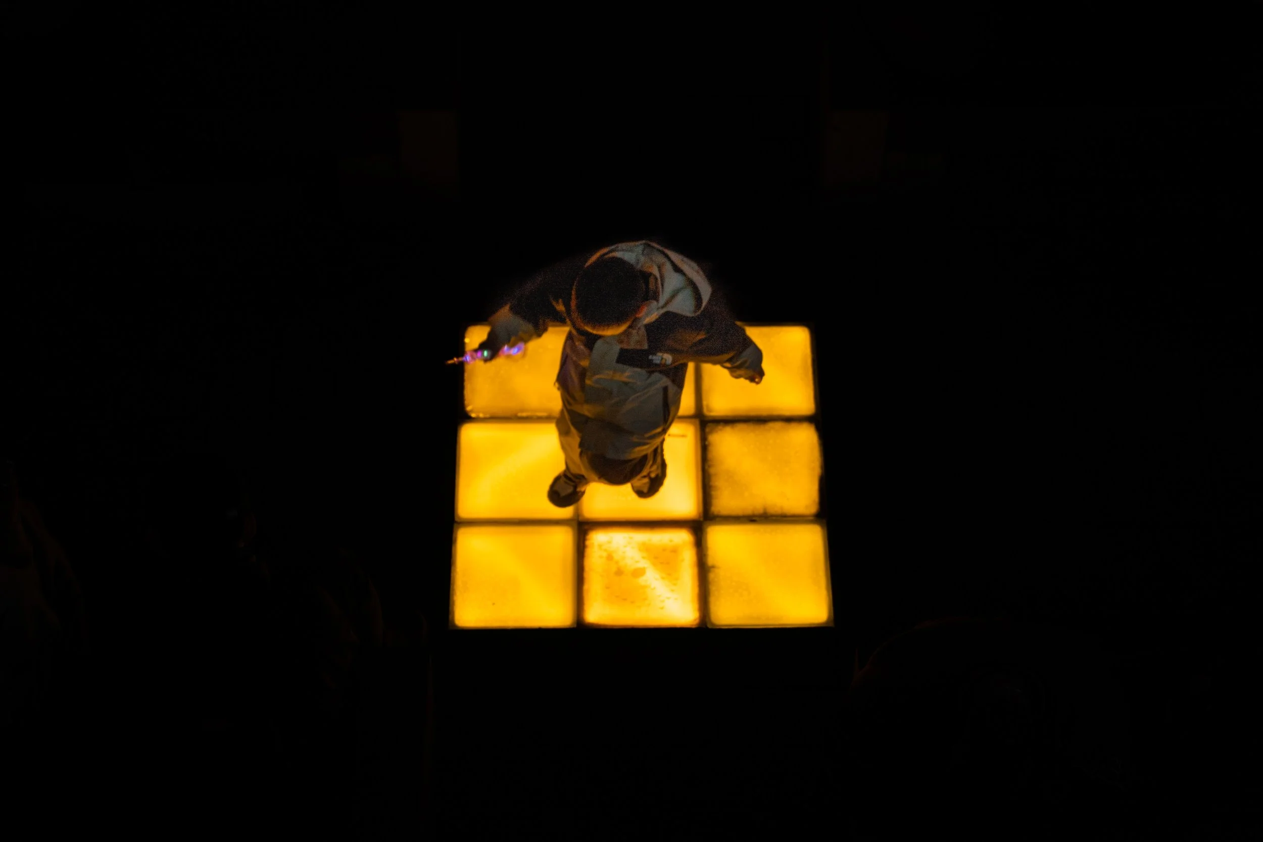 Overhead view of a person standing on illuminated yellow tiles in a dark space, holding a glowing object in one hand.