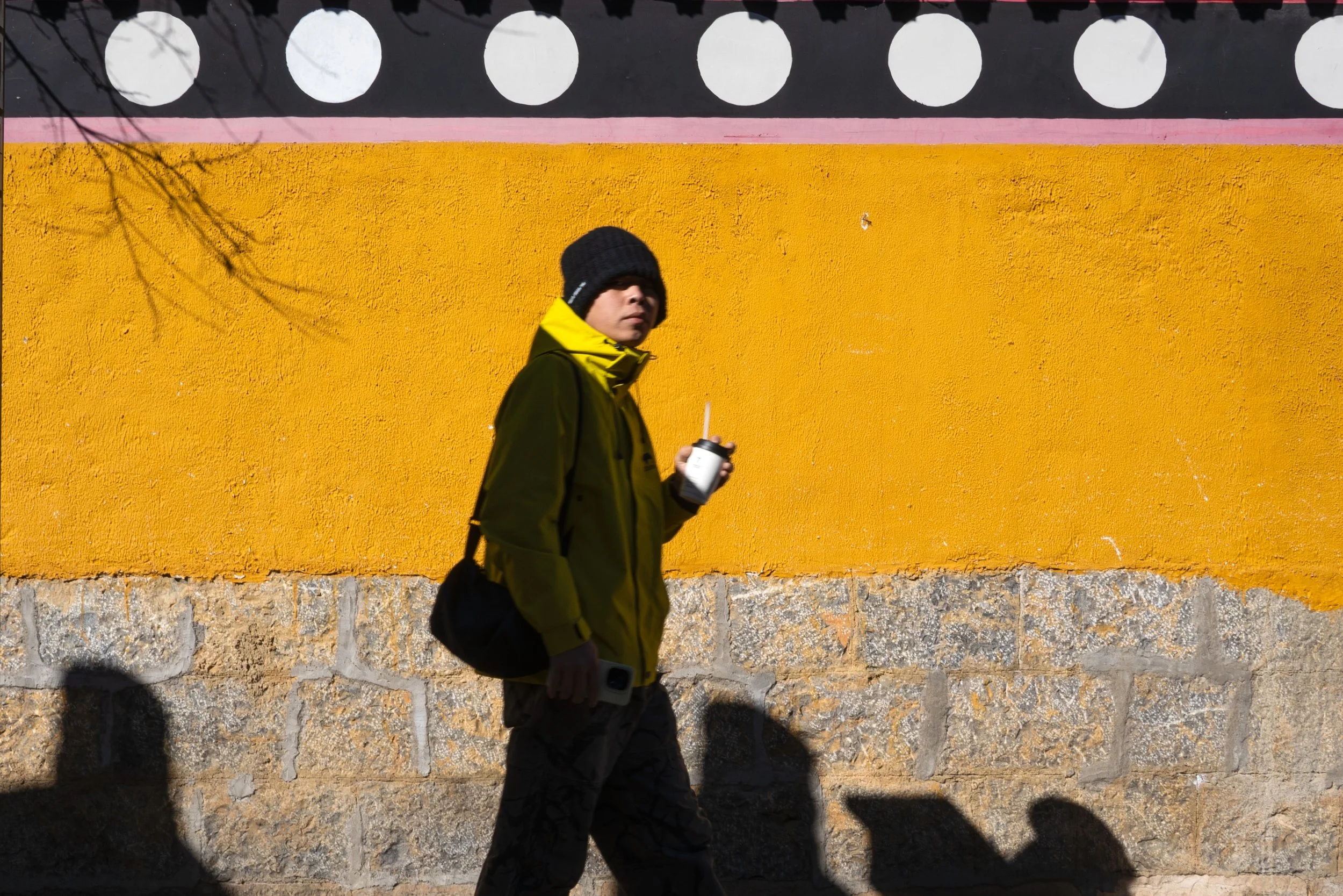 A person walking along a colorful yellow and pink wall, wearing a black beanie and dark clothing, holding a drink and a camera.