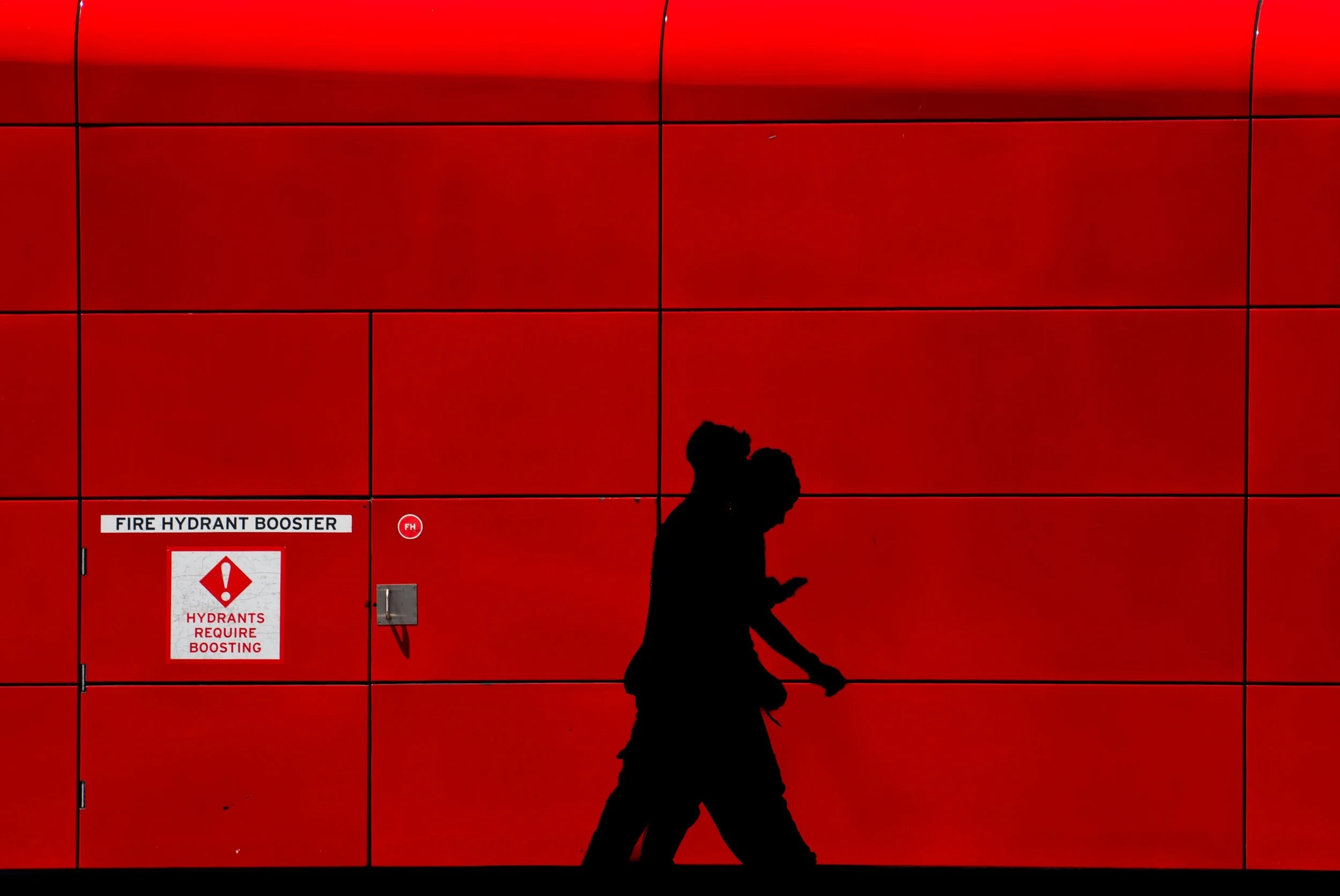 Silhouette of a person walking while looking at a phone in front of a red fire station wall with emergency signage.
