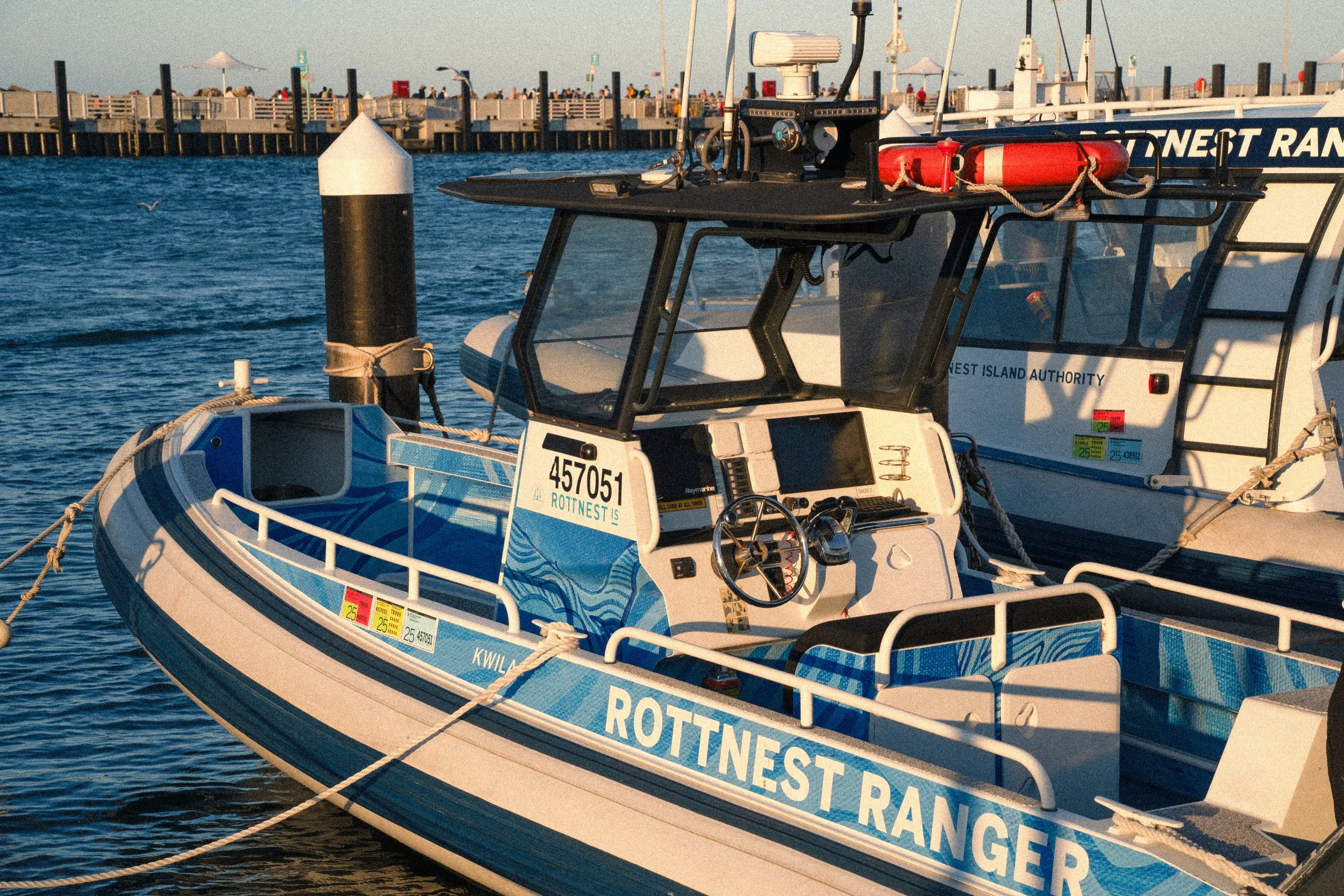 A rescue boat with blue and white colors, labeled 'ROTTNEST RANGER,' is docked at a marina. The boat has a steering wheel, navigational equipment, and safety gear on board.