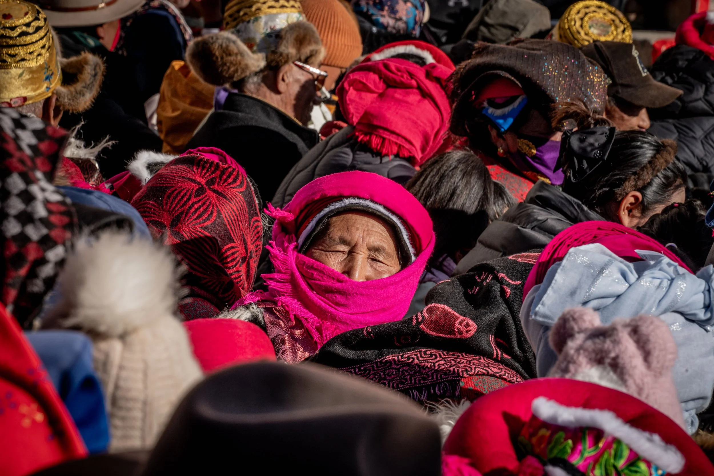 A group of people wearing warm clothing, hats, and scarves, gathered closely together, possibly outdoors in cold weather.