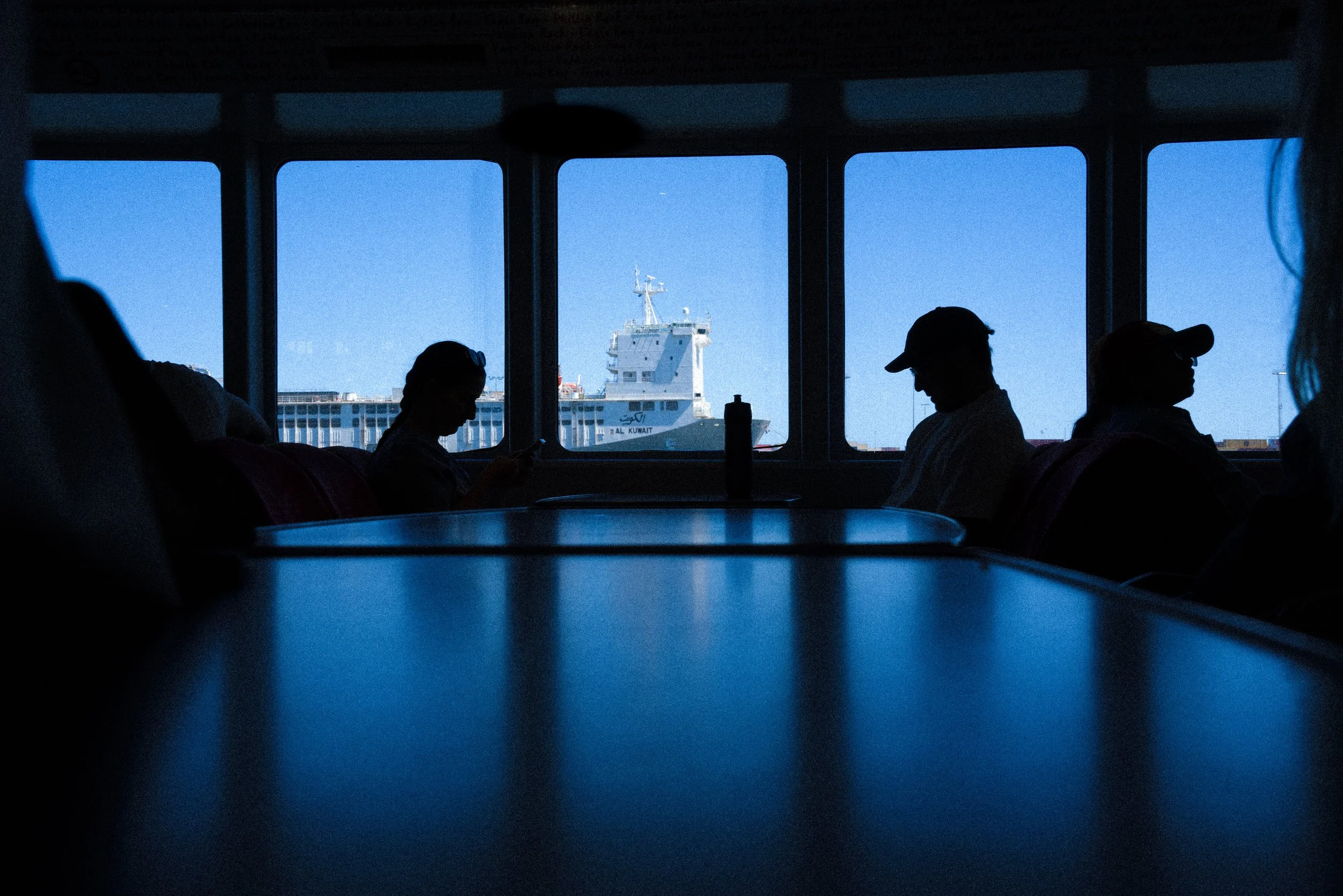 Silhouetted passengers sitting inside an airport waiting area or waiting lounge, with a large window showing a ship anchored outside, labeled 'Al Kuwait,' against a clear blue sky.