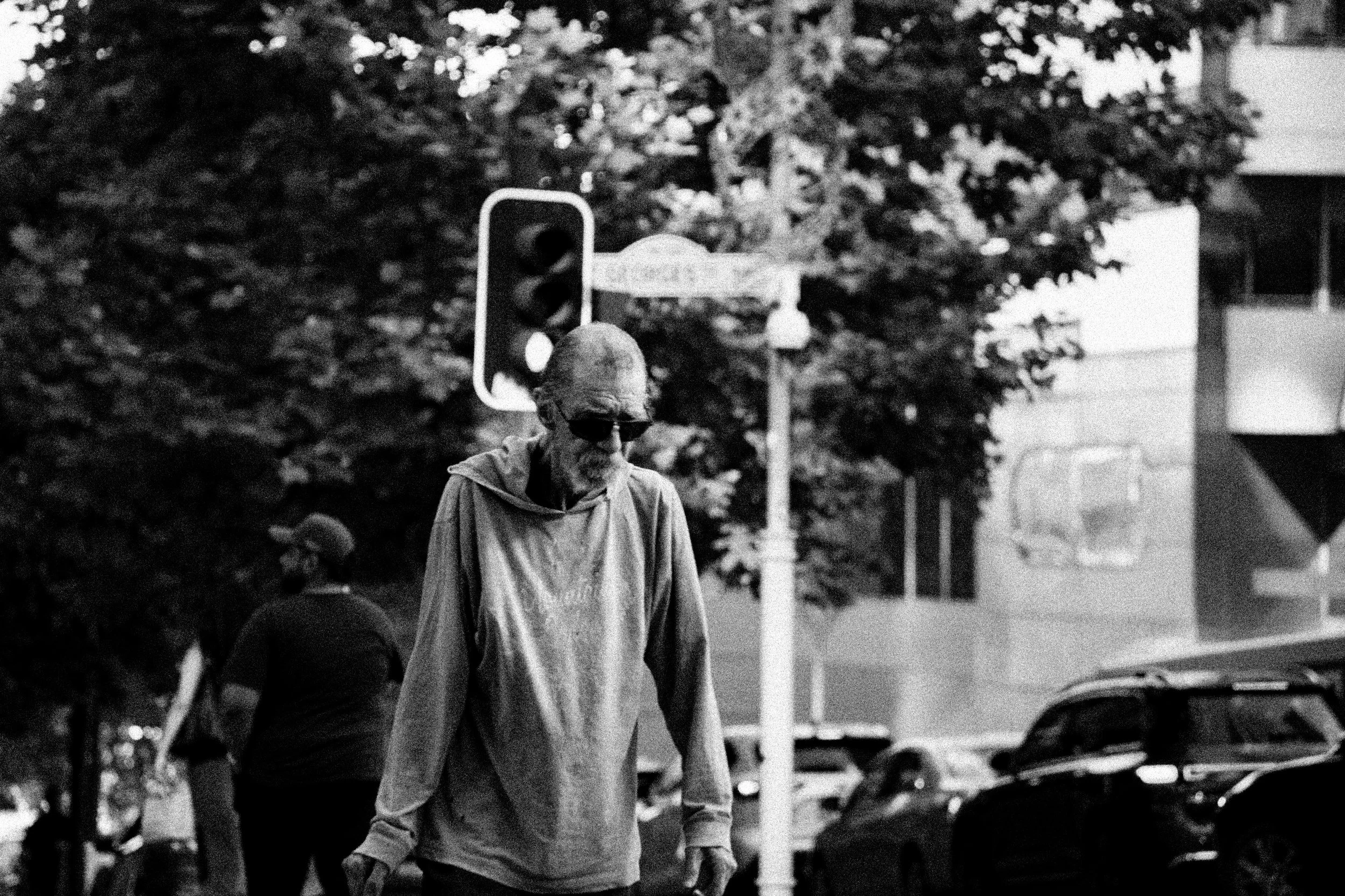 A black and white photo of two people walking outdoors. One person in the foreground with sunglasses and a hoodie, looking down, and another person in the background. There are trees, a traffic light, and parked cars.