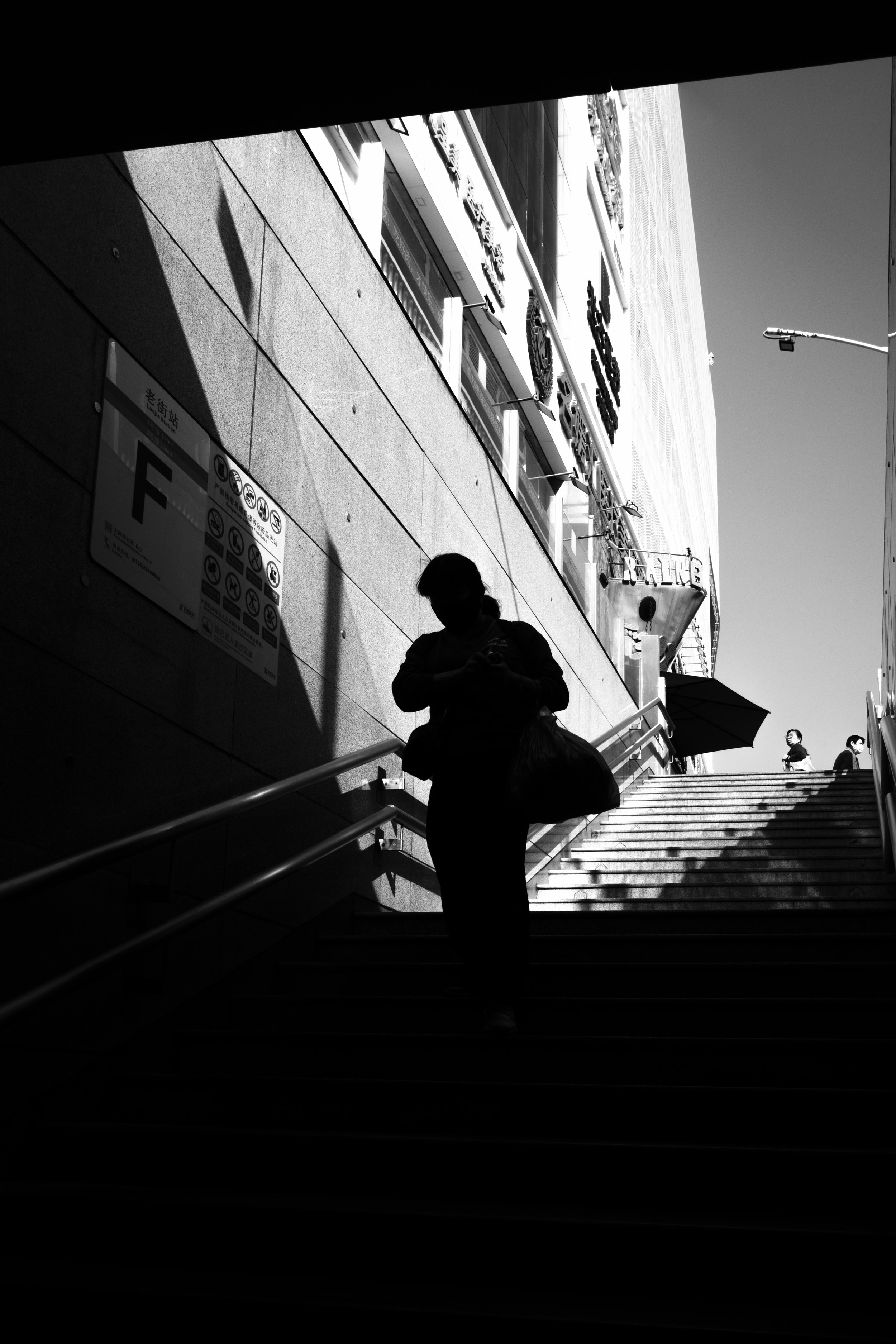 Silhouette of a person walking down stairs in a city environment, with shadows and sunlight creating high contrast. There is a building exterior and signage visible, with some people walking in the background.