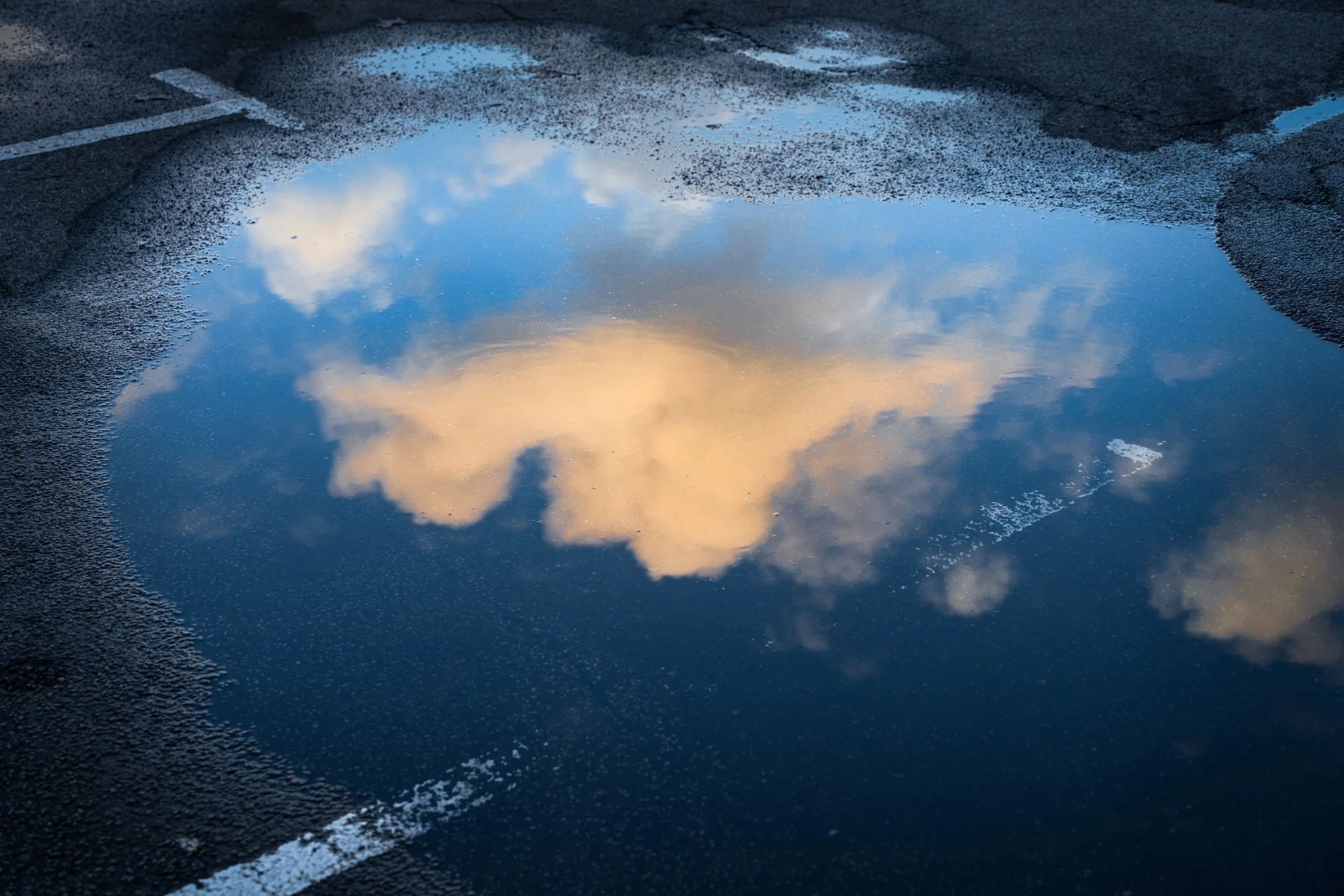 Reflection of a cloudy sky with orange and blue hues in a puddle on an asphalt surface.