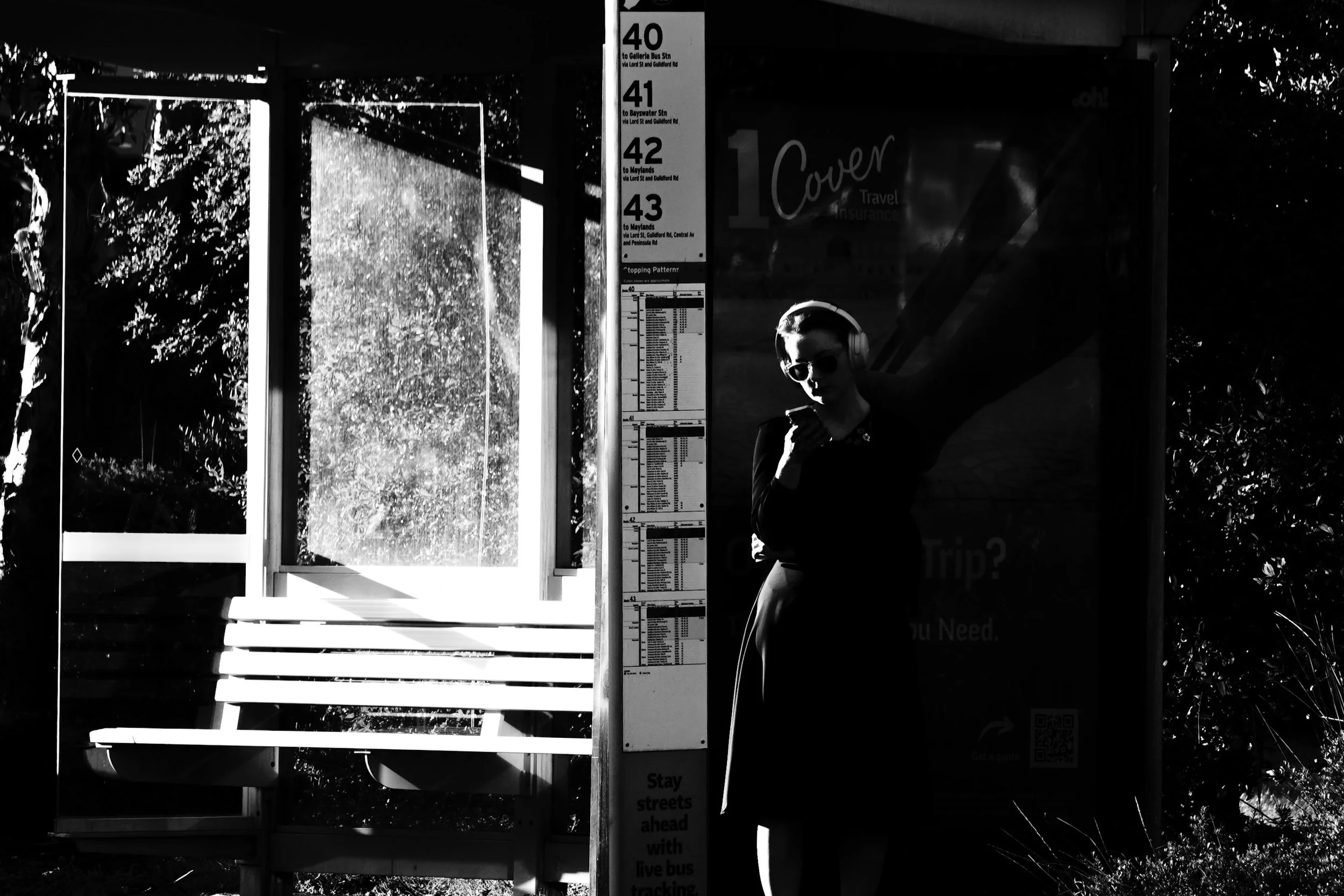 A woman standing at a bus stop, holding a phone, wearing sunglasses and headphones, with sunlight casting shadows on her and the surrounding area, including a bench and bus schedules.
