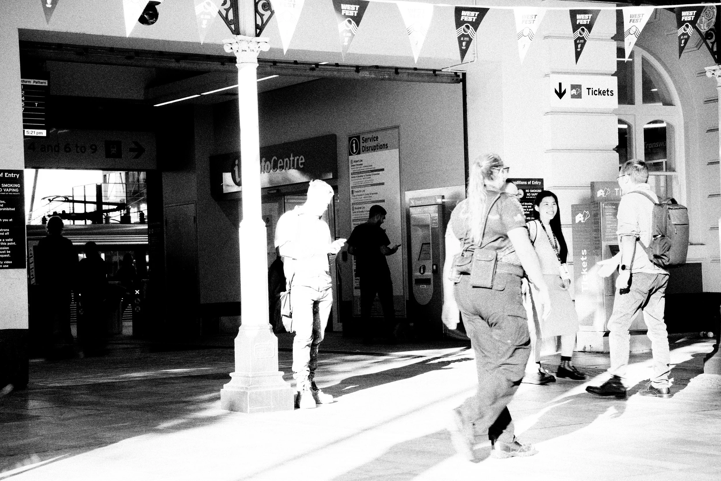 People walking and standing inside a busy train station with signs for tickets, info center, and platform information, in black and white.