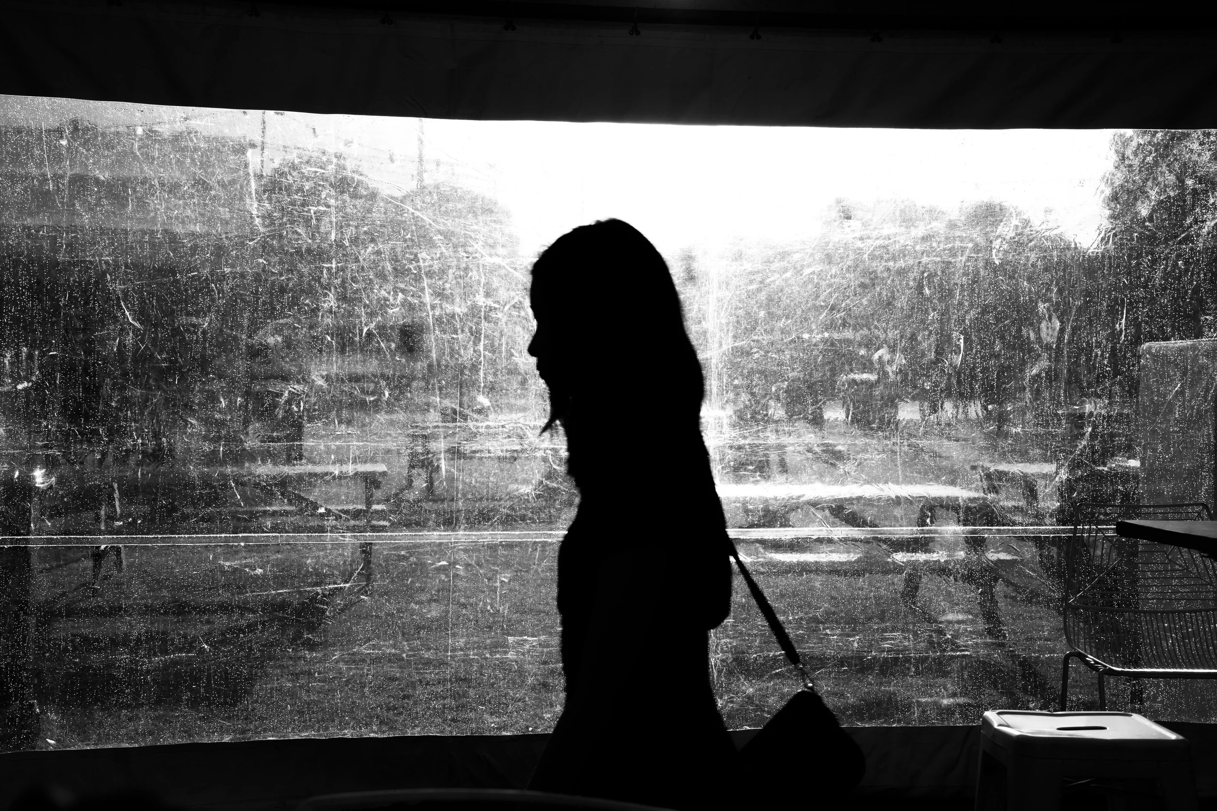 Silhouette of a woman walking past a rain-covered window with cars and trees outside.