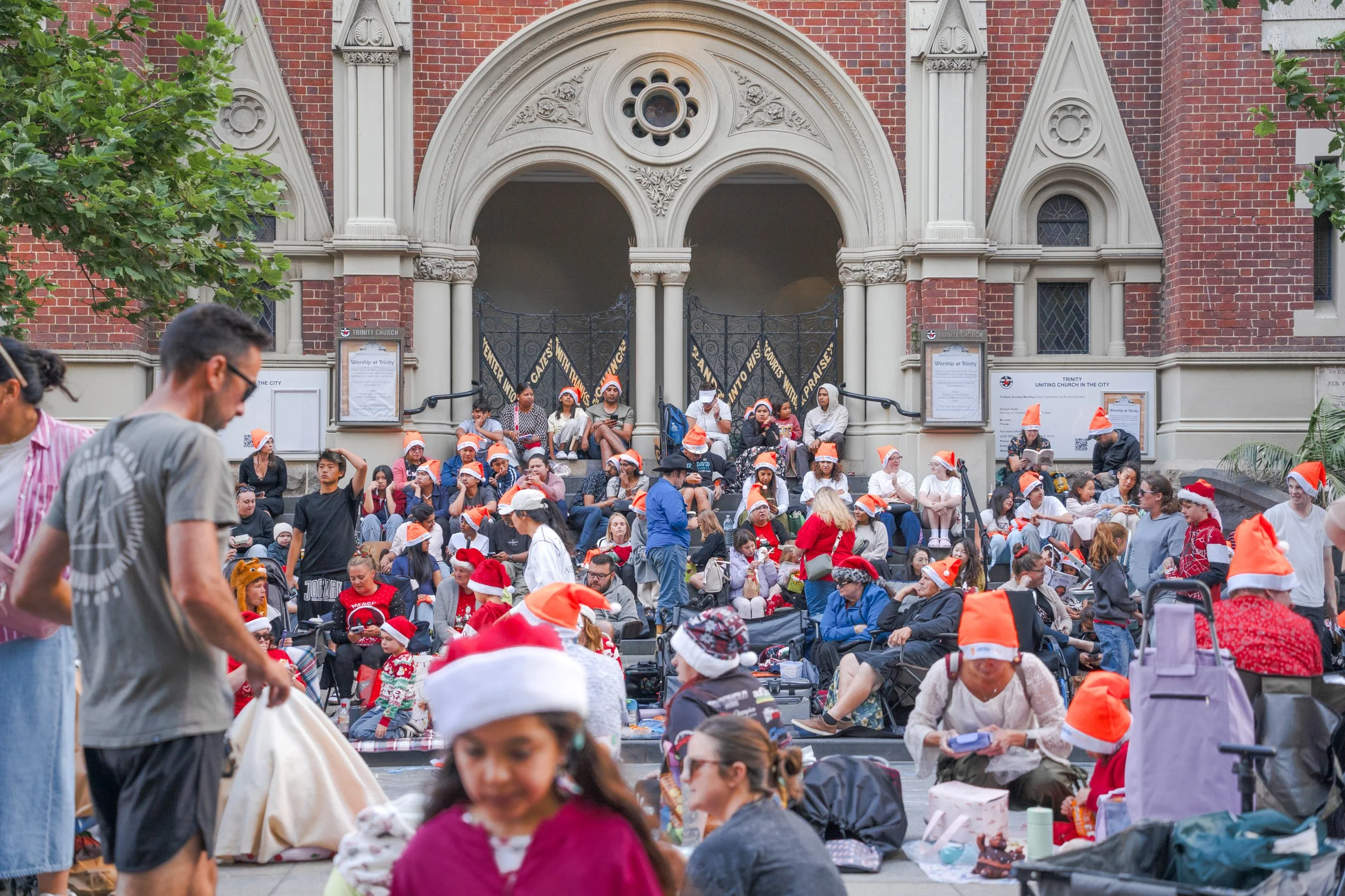 People gathered outside a church wearing red and white Santa hats, many sitting on steps and on the ground, with some standing and moving around. The church has a red brick facade with ornate stone entrance and black wrought iron gates.