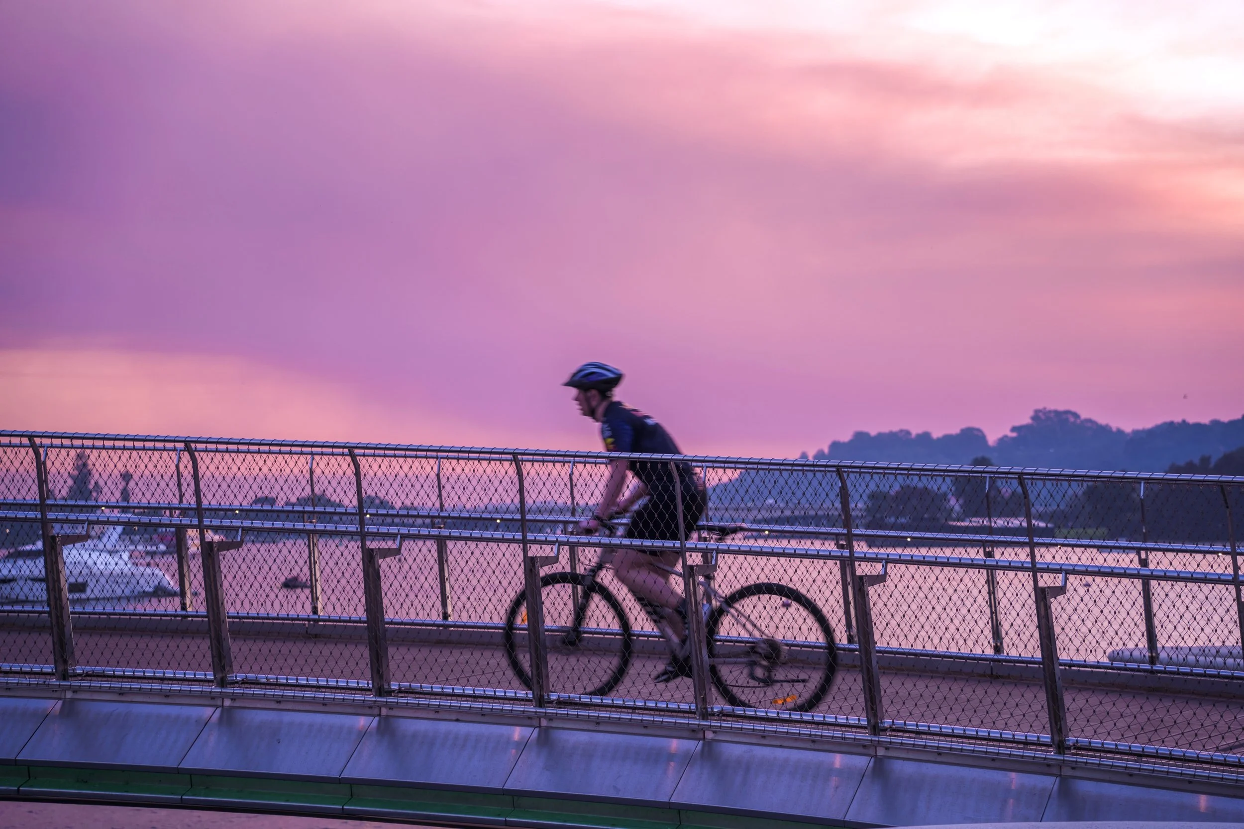 A person riding a bicycle on a curved bridge or pathway with a safety railing during sunset, with a sky painted in pink and purple hues and water and boats visible in the background.