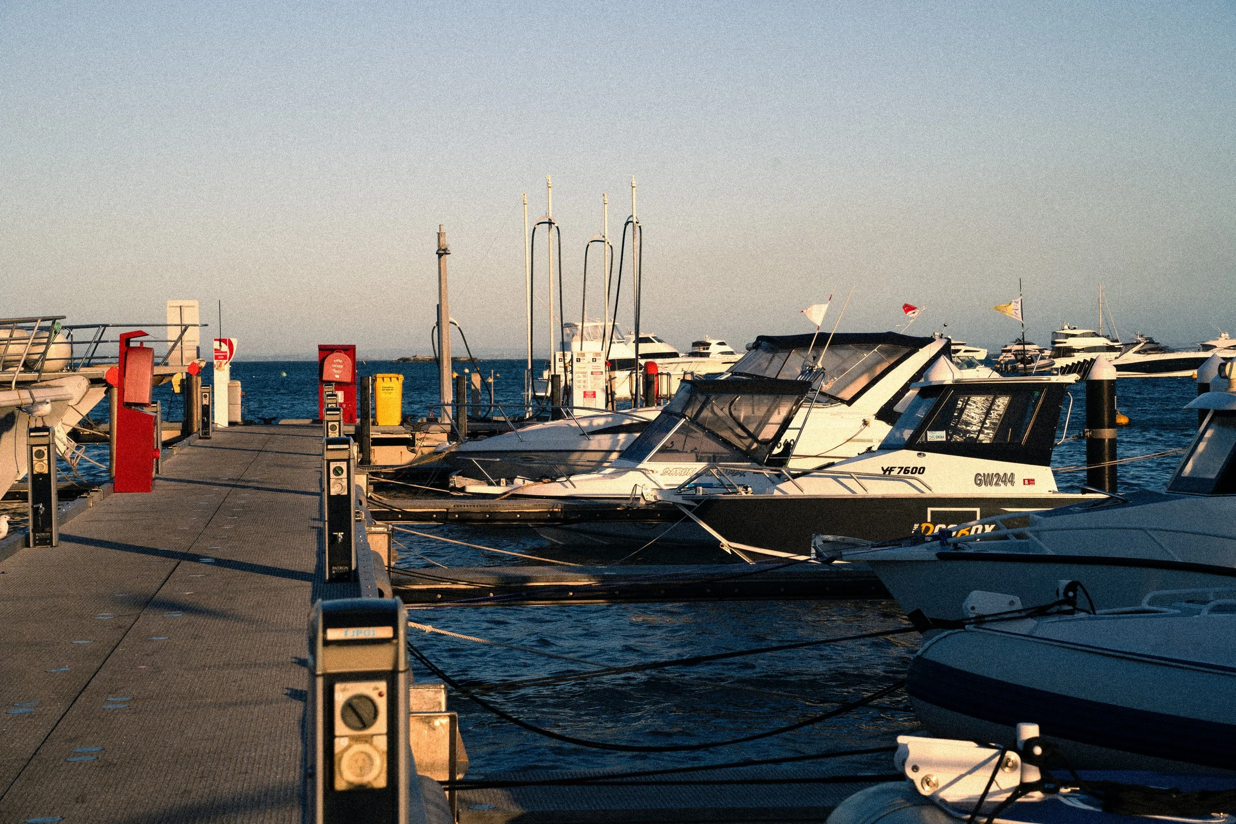 Marina with boats docked at a pier, calm water, clear sky, and distant yachts.