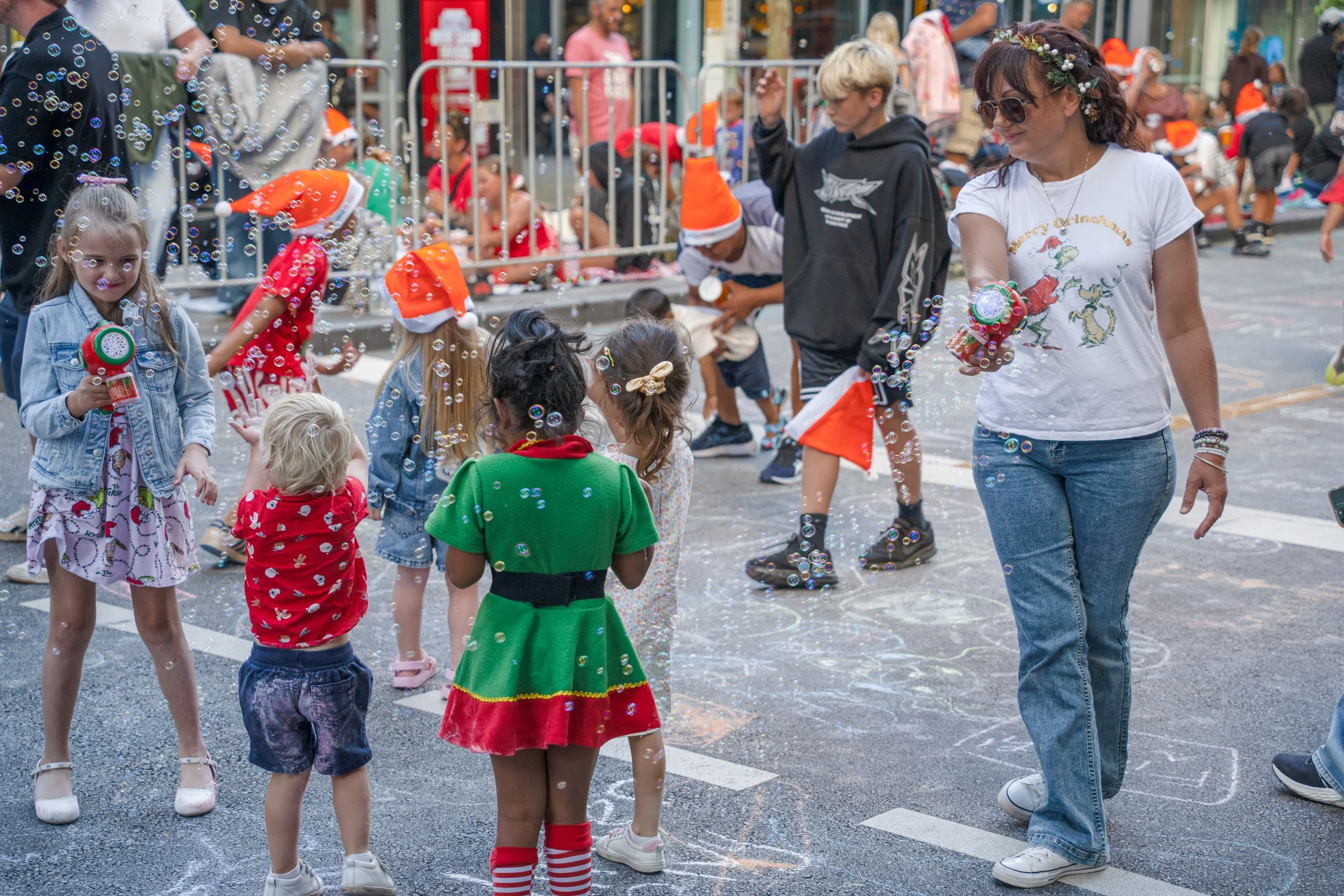 People celebrating Christmas outdoors, with children and adults playing with bubbles. Some children are wearing Christmas-themed clothing and Santa hats, while others are casually dressed. One woman is holding a bubble-blowing toy, and chalk drawings