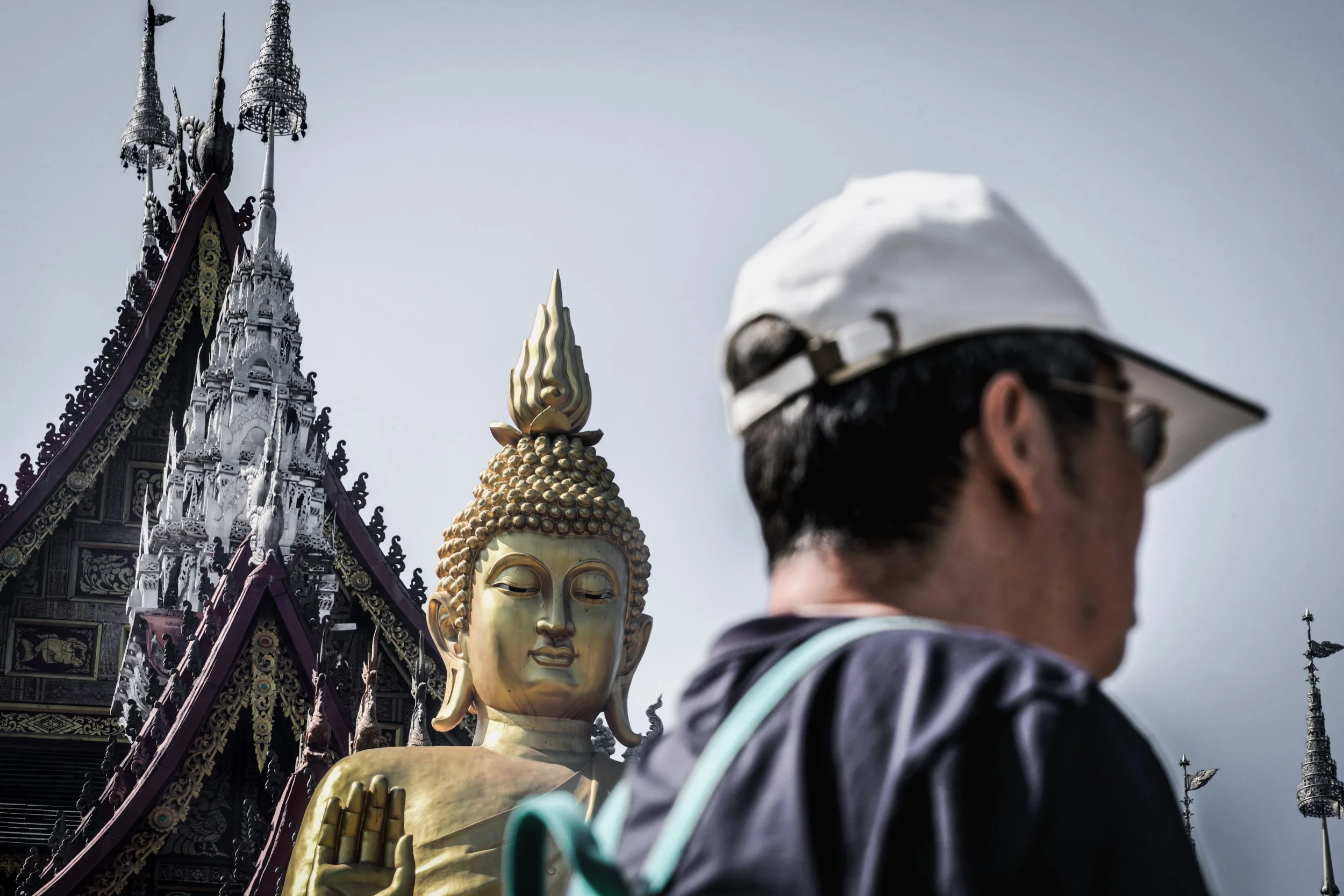 A person wearing a white cap and sunglasses in front of a large golden Buddha statue and ornate temple in Thailand.