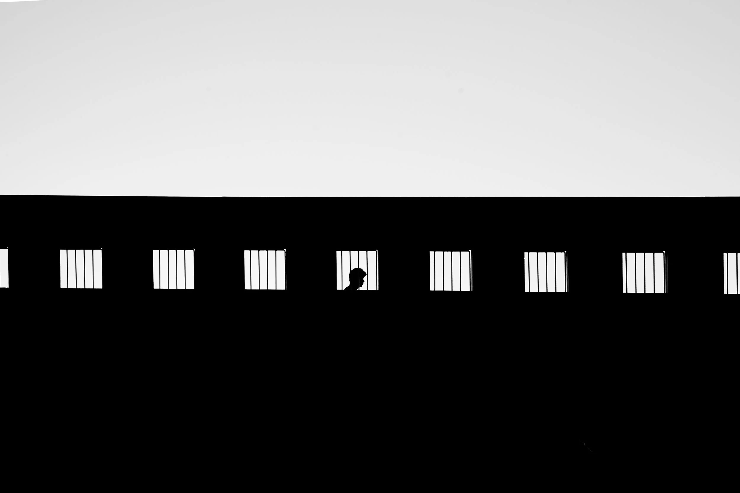 Silhouette of a person behind bars, viewed through a row of barred windows on a building, in black and white.