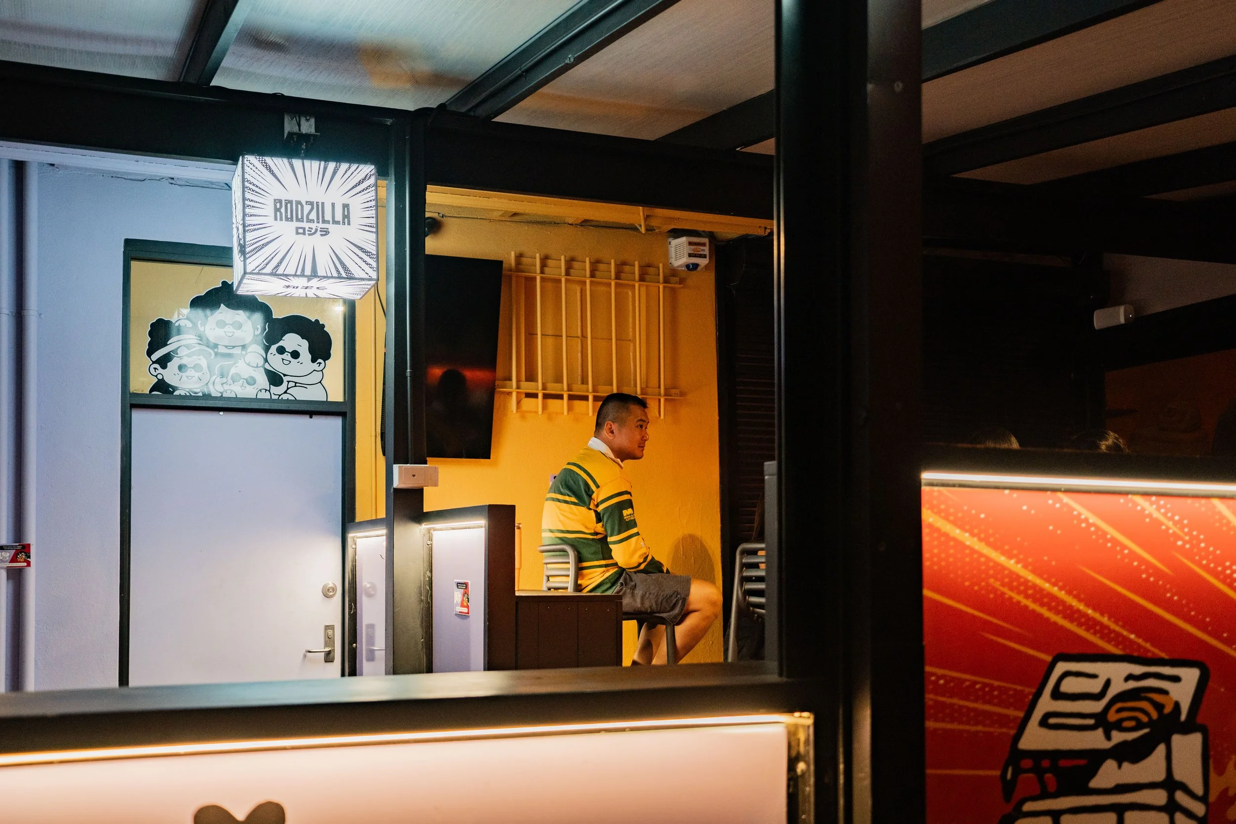 A man sitting in a small, brightly lit room with yellow walls, wearing a green and yellow striped shirt and gray shorts, looking to his right. The room has a yellow wall with a small wooden rack and a television mounted on the wall. In the foreground