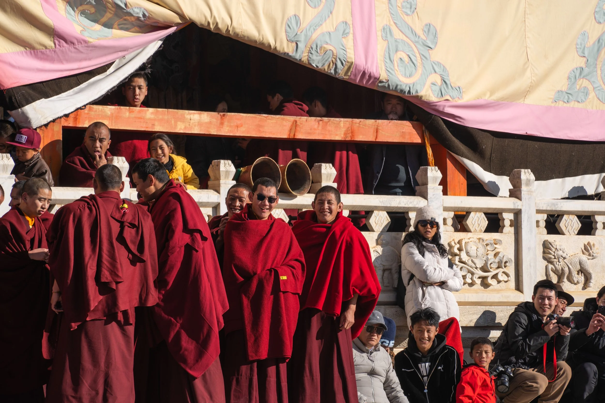 Group of monks and tourists gathered on a decorative stone terrace, some smiling and some taking photos, under a colorful canopy.