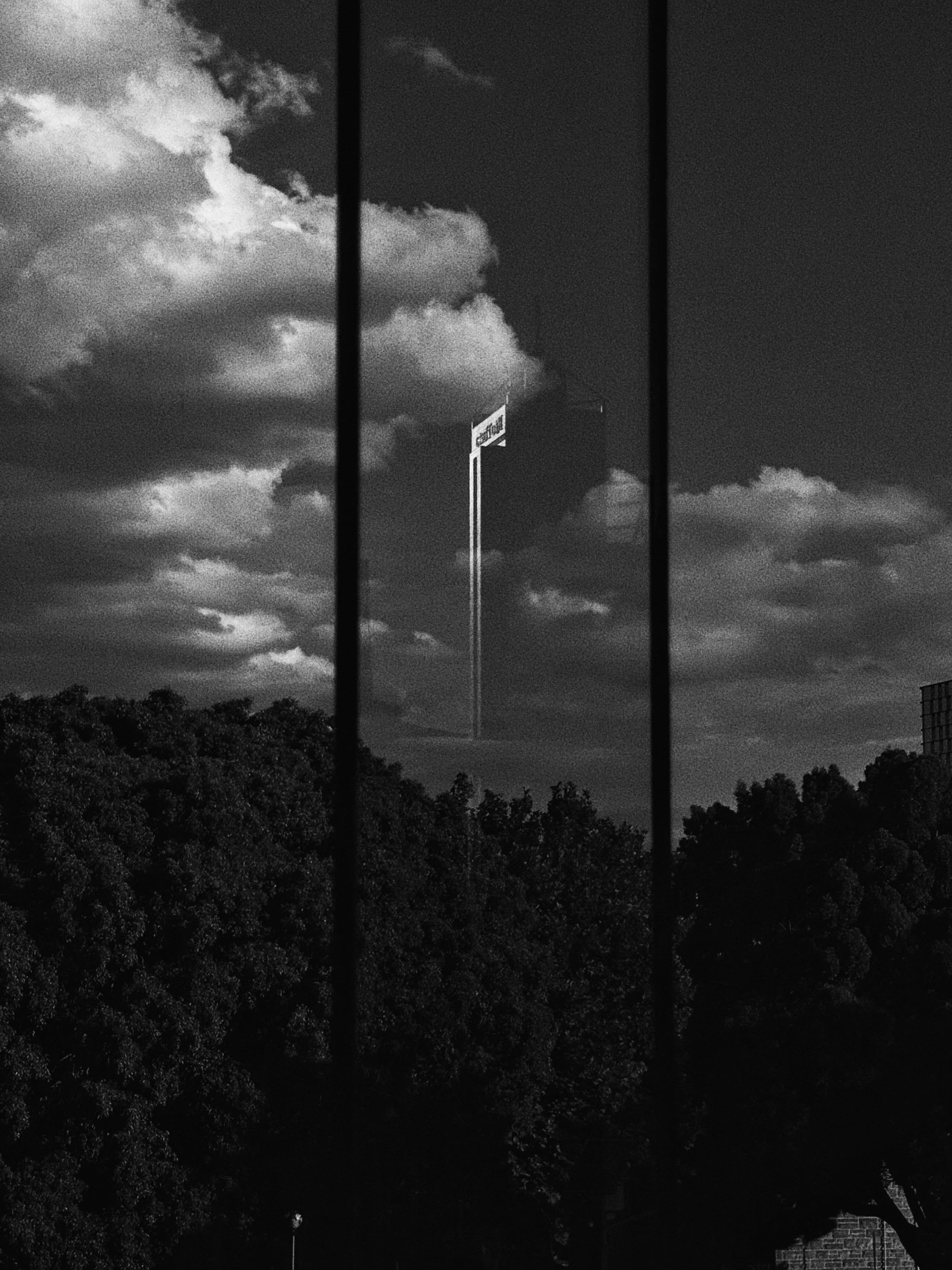 Black and white photo of a cityscape viewed through window bars, with a tall billboard in the distance under a cloudy sky.