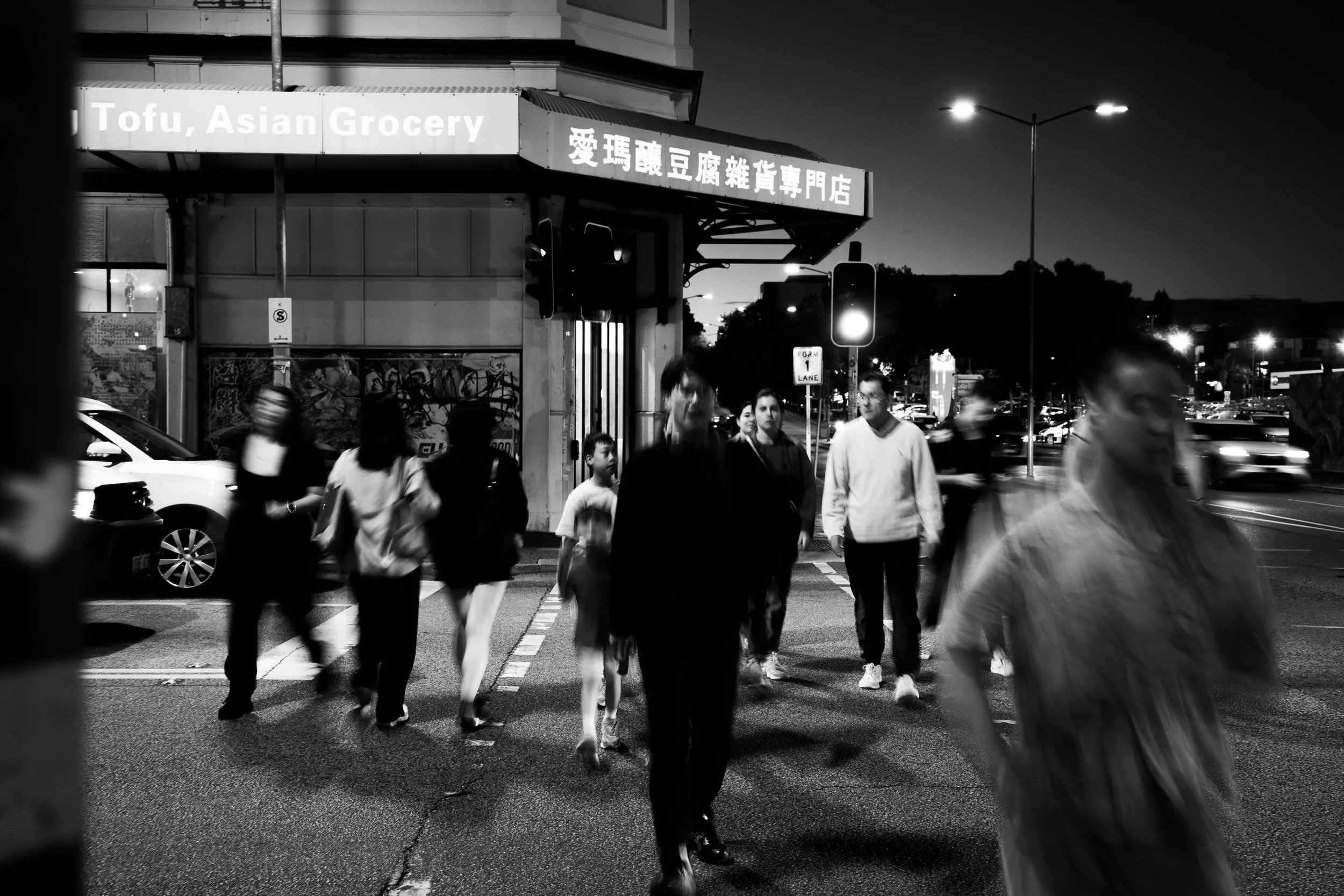 Black and white photo of people crossing a street at night, with a sign for an Asian grocery store that sells tofu, in the background.